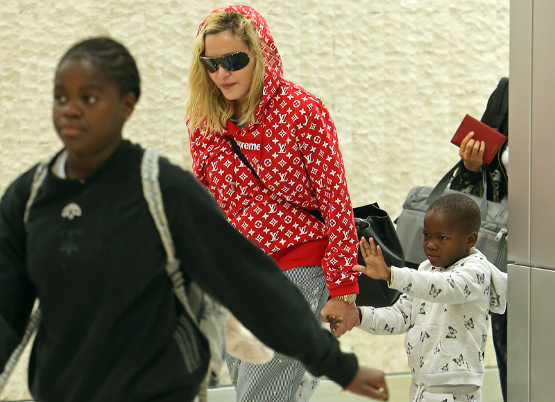Madonna rocks a red supreme louis vuitton hoodie while touching down at JFK airport in New York holding her twins hands Estere and Stelle while Lourdes, David, and Mercy in tow after celebrating her 59th birthday in Italy.