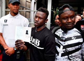 Malian migrant Mamoudou Gassama (C) flanked by his older brother (R) holds his temporary residence permit after receiving it at the Prefecture of Bobigny, northeast of Paris on May 29, 2018, one day after he was honored by the French President for scaling an apartment building to save a 4-year-old child dangling from a fourth-floor balcony. - Two days after his daring rescue -- viewed millions of times online -- Mamoudou Gassama, 22-year old, nicknamed "Spiderman" by French media for his astonishing climbing ability and feted as a hero, was offered by French President citizenship, and a job with the fire service.