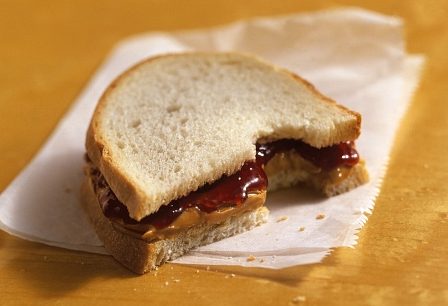 Half a peanut butter and jelly sandwich with a bite taken out of it sitting on a piece of parchment paper sitting on a wooden surface.
