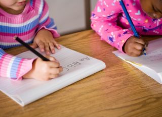 Two girls (5-7) writing at desks in classroom, mid section - stock photo