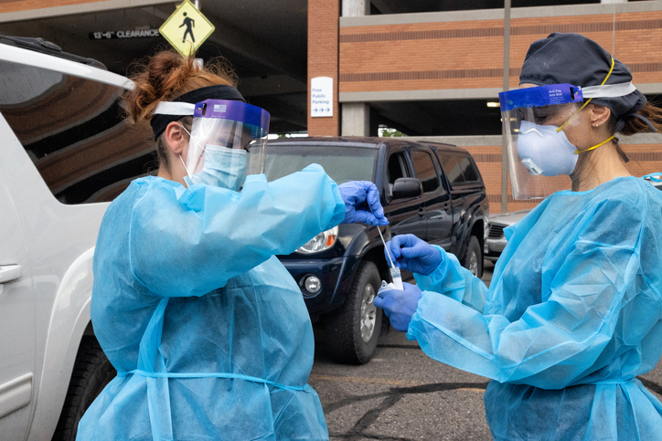 A Female Nurse Wearing a Gown, Surgical Face Mask, Gloves, and a Face Shield Places a Used Cotton Swab into a Test Tube Held by Another Nurse in a Drive-Up COVID-19 Testing Line Outside a Medical Clinic/Hospital Outdoors