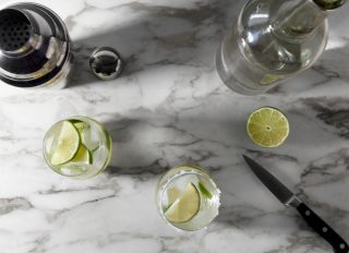 Two margaritas with cocktail shaker, tequila bottle, and limes on marble