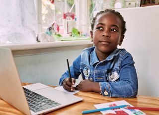 A young African girl in her room holding a pencil and paper in front of her laptop, contemplating something.