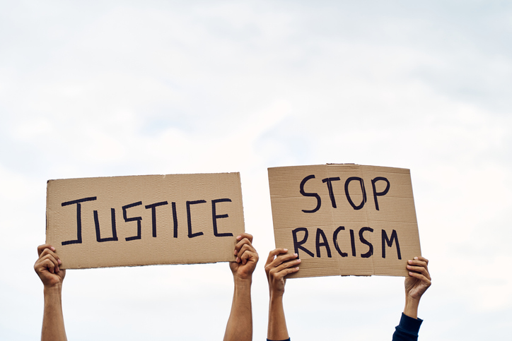Unrecognizable man and woman holding protest banners with messages to STOP RACISM AND JUSTICE, with the sky in the background.