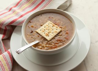 Bowl of vegetarian barley soup with a salt cracker garnish in a white bowl with saucer on a white marble table