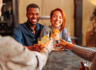 Young African-American man and Hispanic woman toasting cocktail glasses with friends in restaurant.