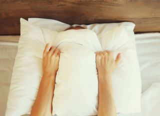Unhealthy sleep, woman suffering from insomnia lying on the bed and holding in hands covering her head with white pillow in bedroom at home