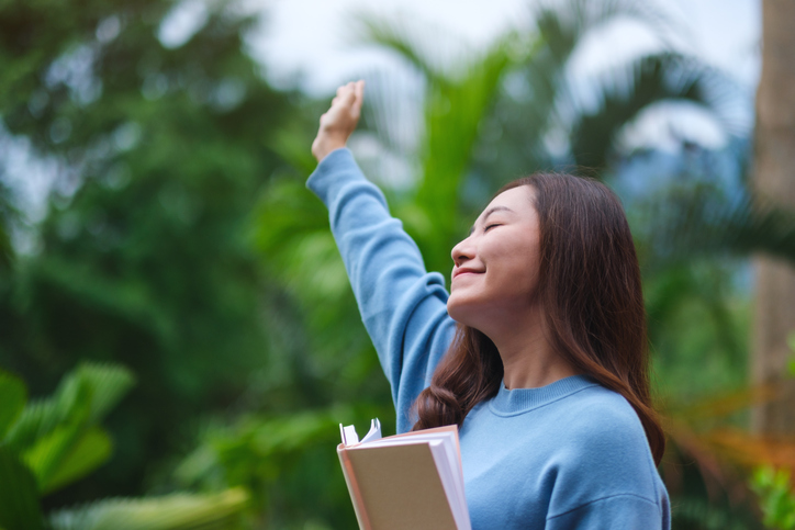 Portrait image of a woman holding a book and raising hand in the park
