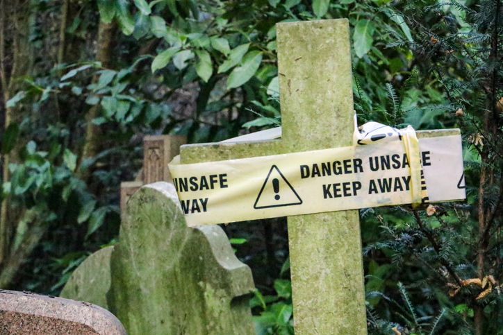 Damaged Victorian stone cross in Highgate Cemetery with danger unsafe keep away sign rapped around it. The Cemetery is one of London's magnificent seven graveyards and is now a unique urban wild area