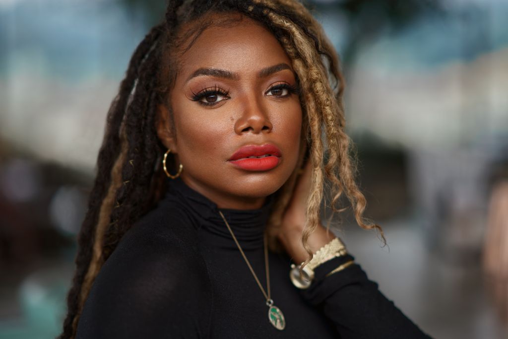 A close-up portrait of a woman with long, curly hair and bold makeup, including red lipstick, looking directly at the camera with a serious expression.