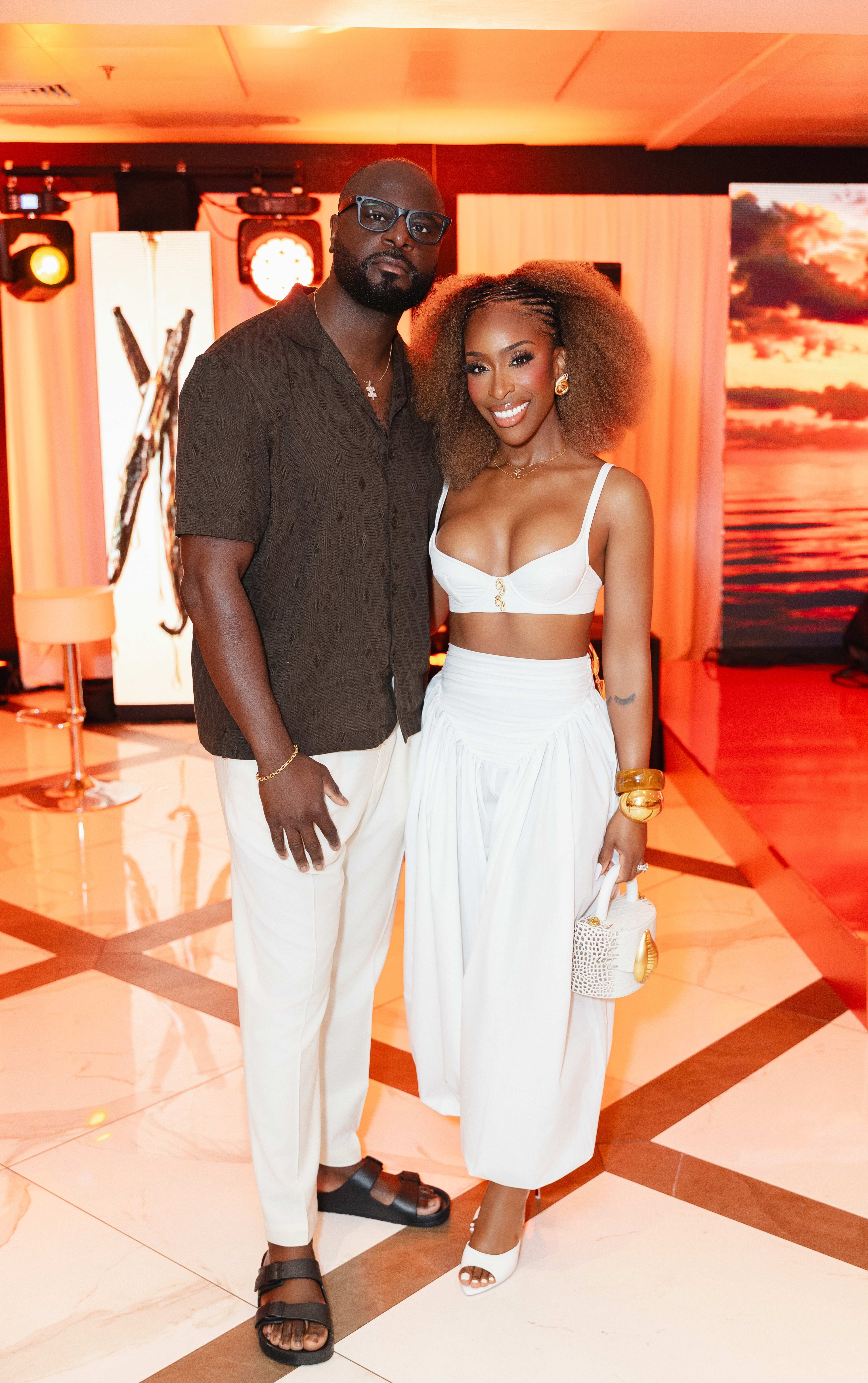 A man and woman posing together in a colorful, illuminated setting. The woman is wearing a white crop top and skirt, and the man is wearing a dark shirt.