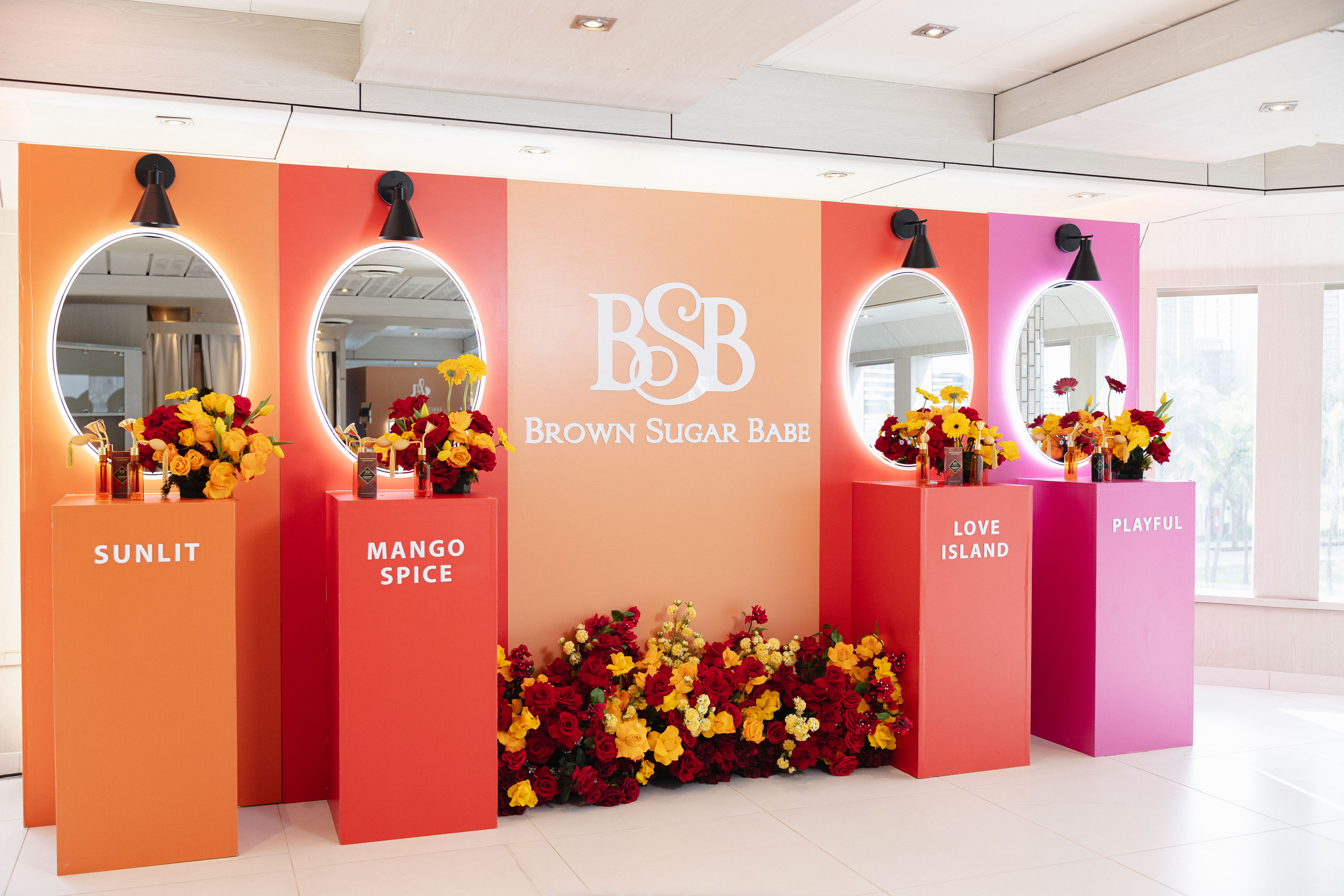 A colorful retail display featuring the Brown Sugar Babe brand, with floral arrangements and mirrored panels showcasing the "Sunlit", "Mango Spice", "Love Island", and "Playful" product lines.