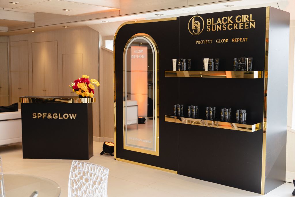 A display stand for Black Girl Sunscreen, featuring shelves of sunscreen products and the brand's logo and slogan "Protect Glow Repeat".