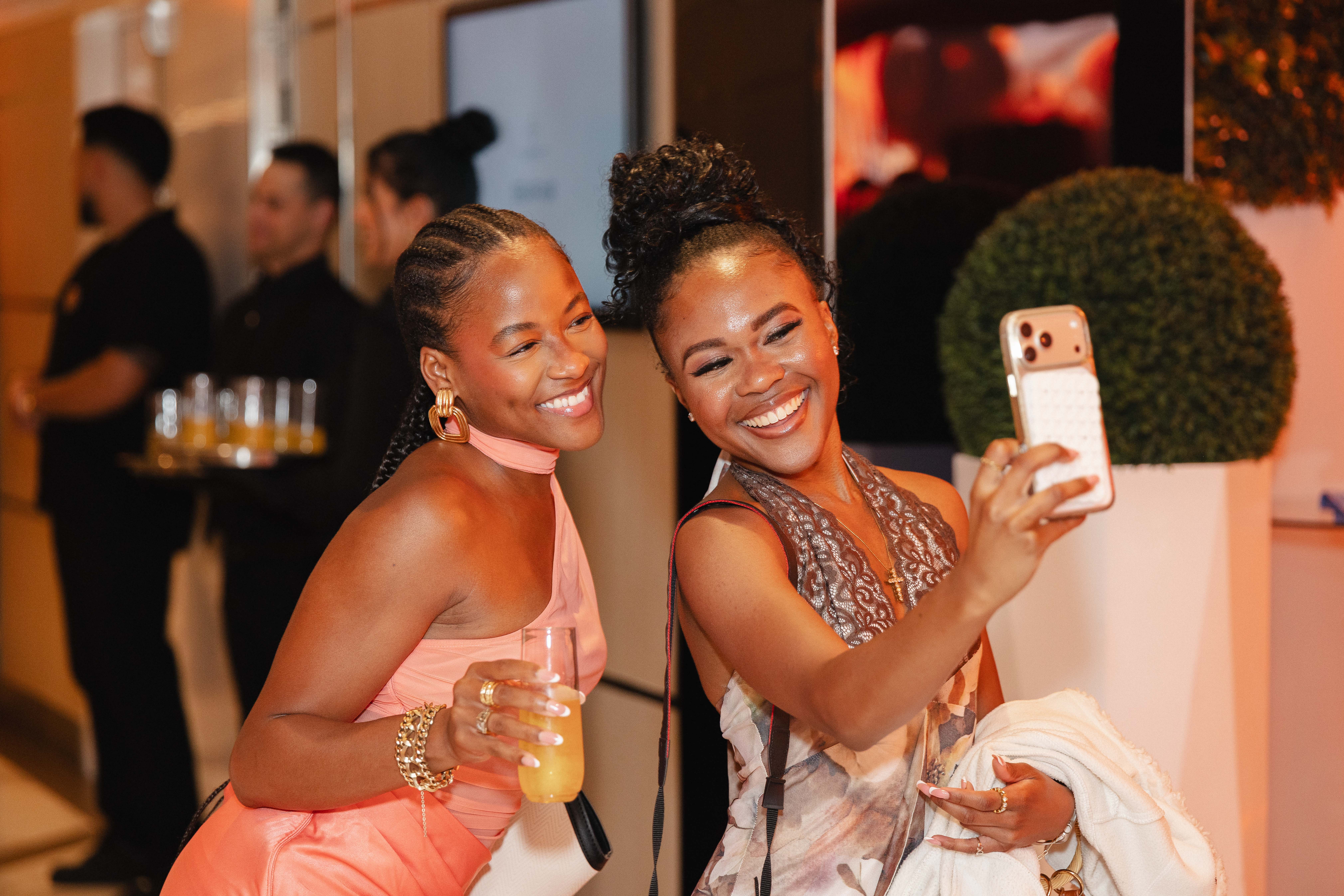 Two smiling Black women taking a selfie at a party, one wearing a pink dress and the other a patterned dress.