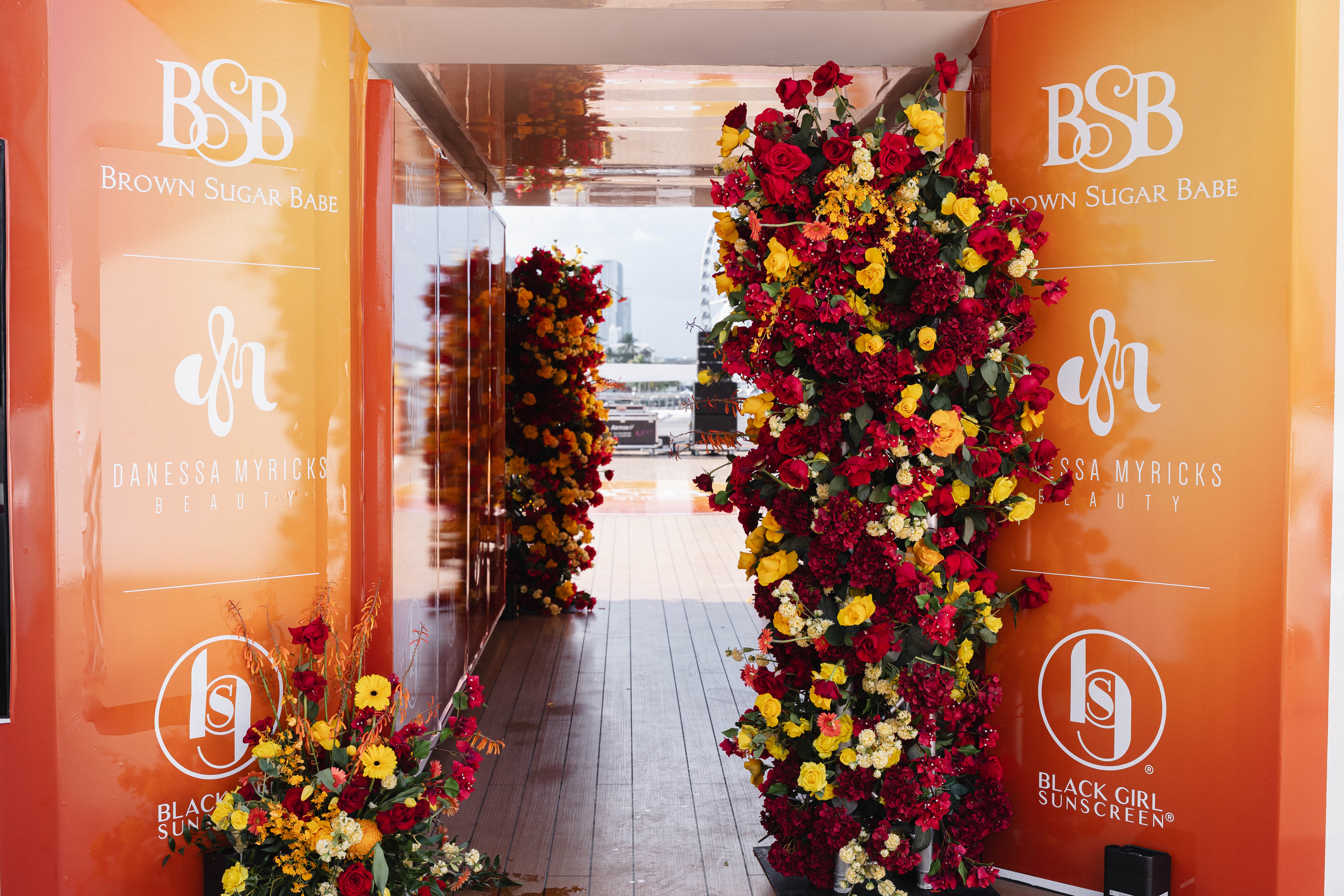Colorful floral display in a hallway with the "Brown Sugar Babe" and "Black Girl Sunscreen" brand logos prominently featured.