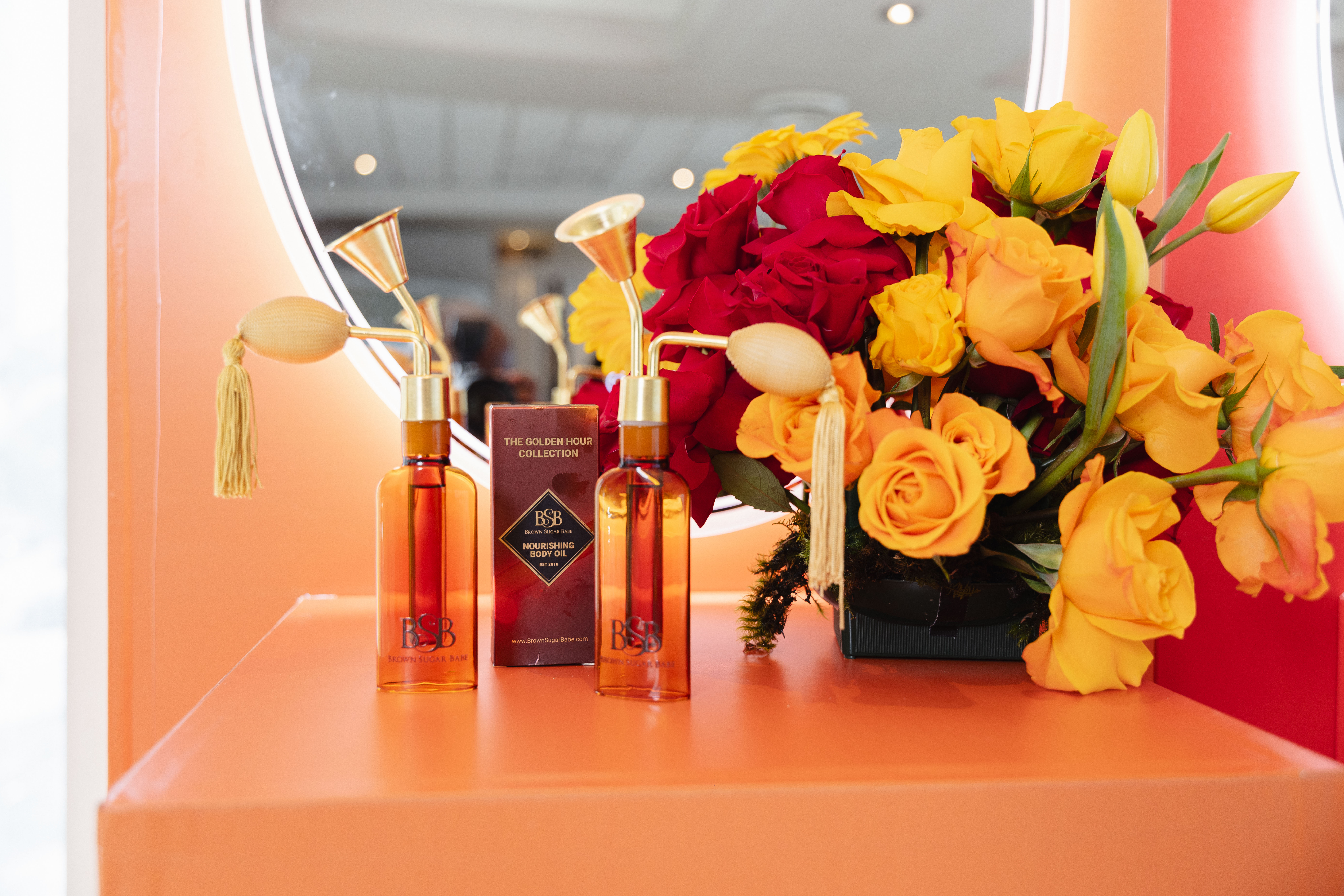 A colorful arrangement of red and yellow roses, tulips, and other flowers in a vase, with bottles of The Golden Hour Collection body oil by B&B.