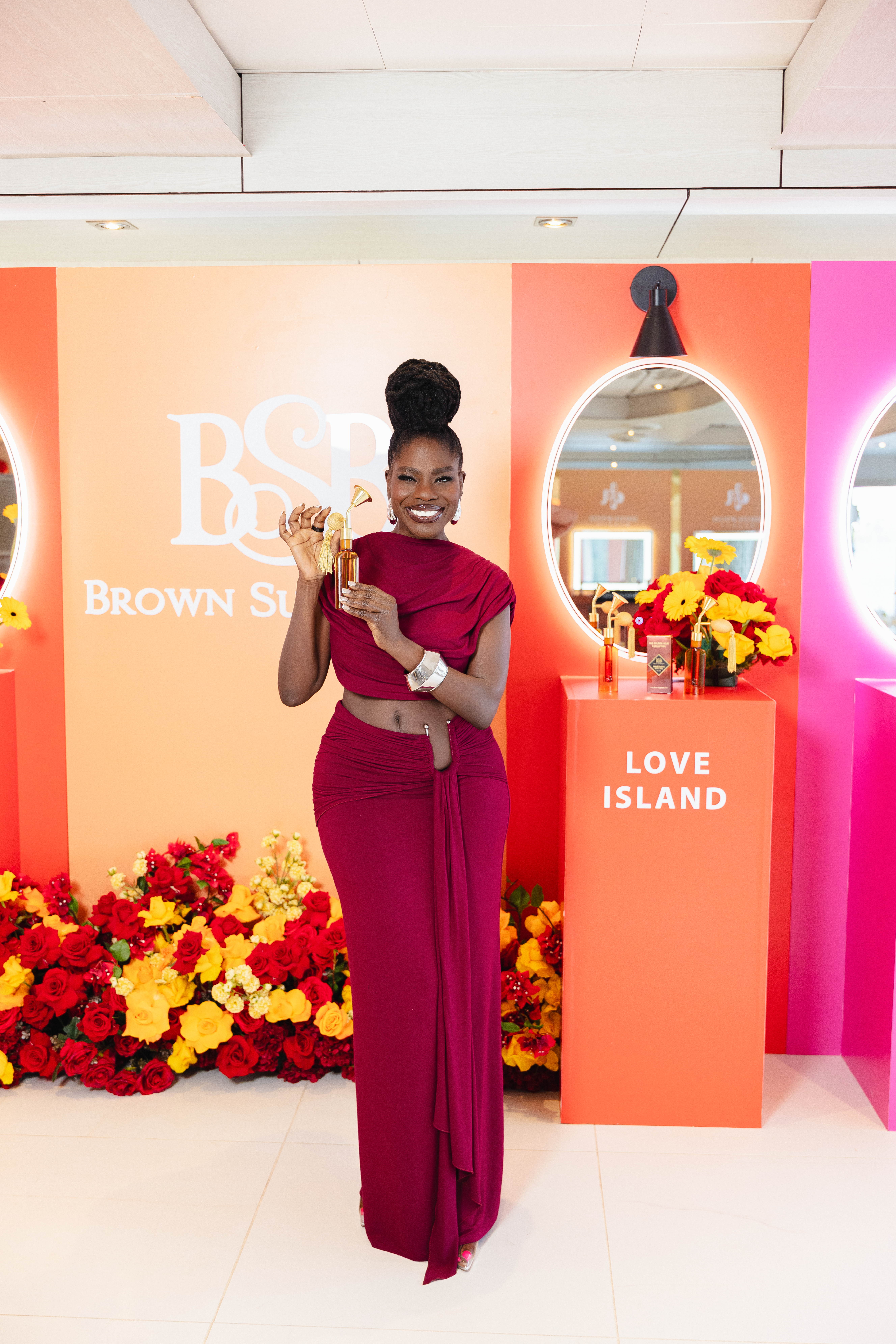 A smiling woman in a red dress poses in front of a colorful display with the "Brown Sugar" and "Love Island" branding.