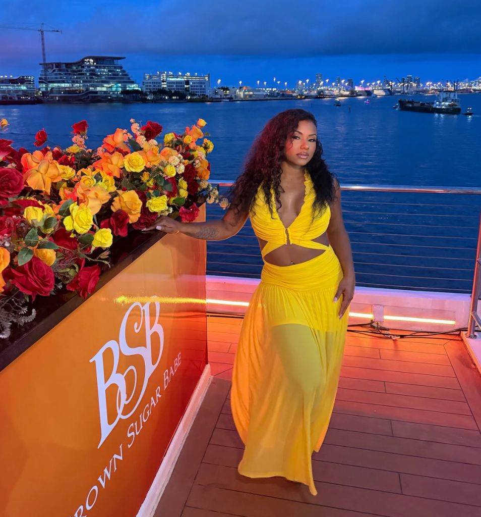 A woman in a yellow outfit stands on a boat deck overlooking a city skyline at night, with a large floral display in the foreground and the text "BSH" visible.