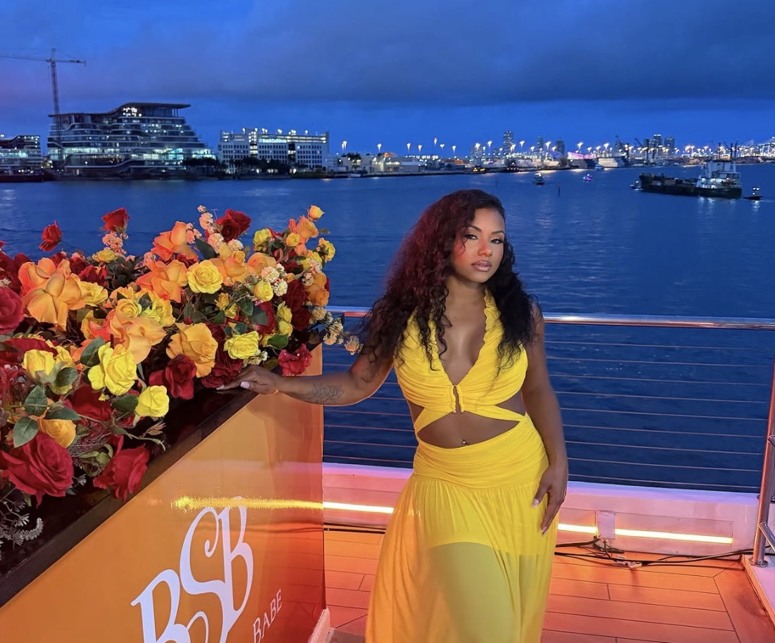 A woman in a yellow outfit stands on a boat deck overlooking a city skyline at night, with a large floral arrangement in the foreground.