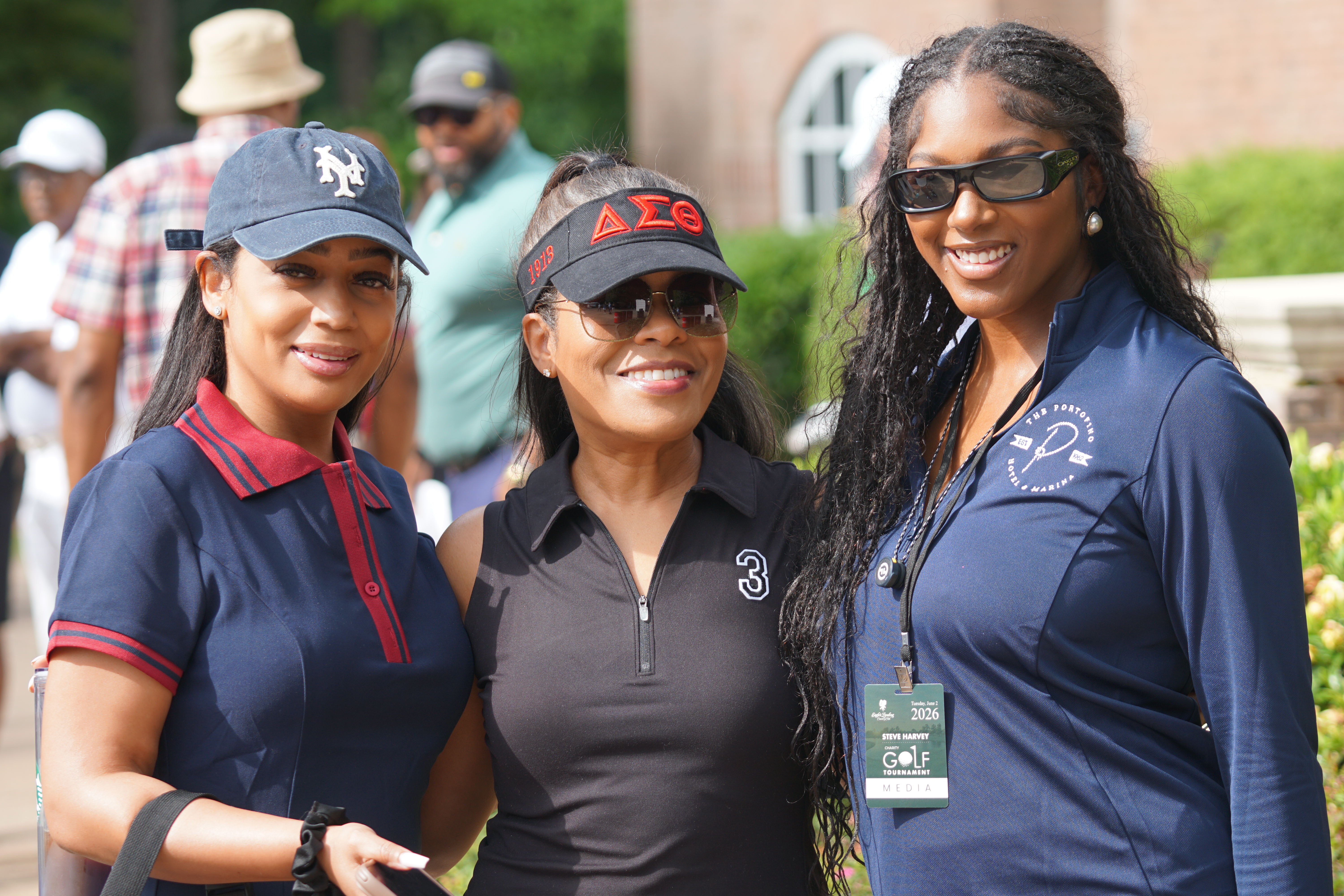 Three smiling women wearing hats and shirts with Greek letters and golf tournament credentials.