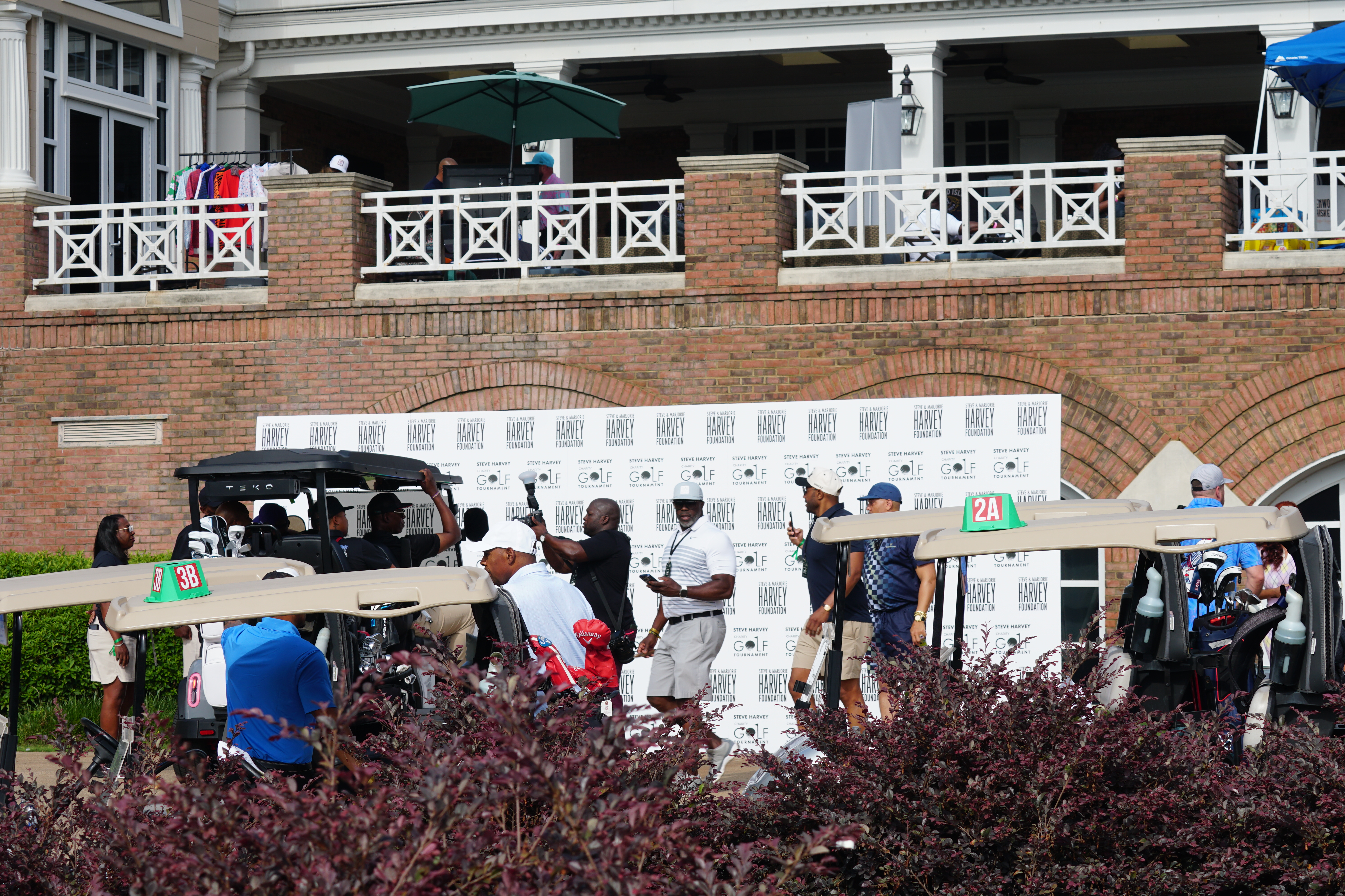 Outdoor seating area with people gathered around golf carts, in front of a brick building with a balcony and "HARLEY" branding.