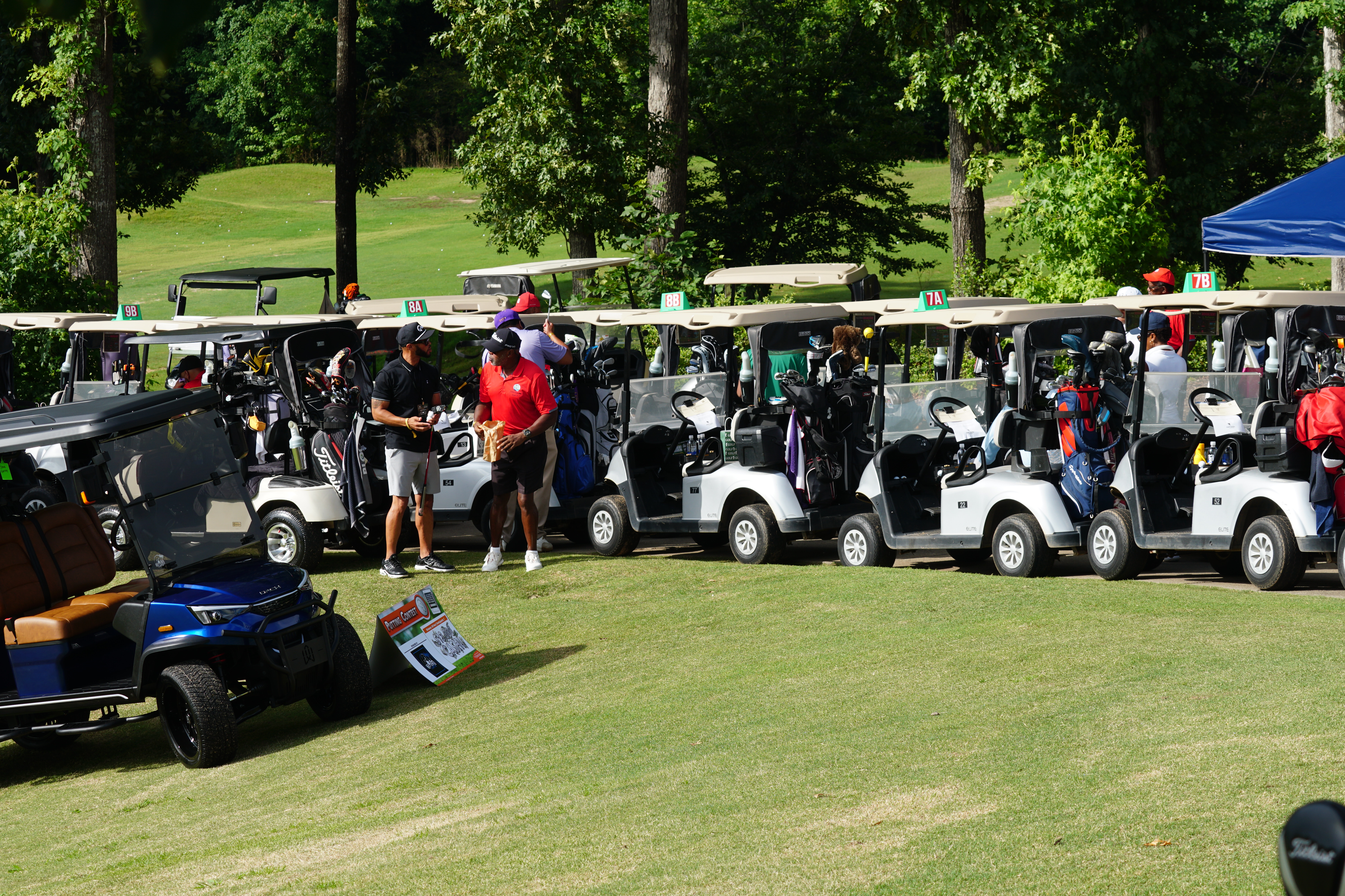 A group of people standing near golf carts on a grassy field surrounded by trees.