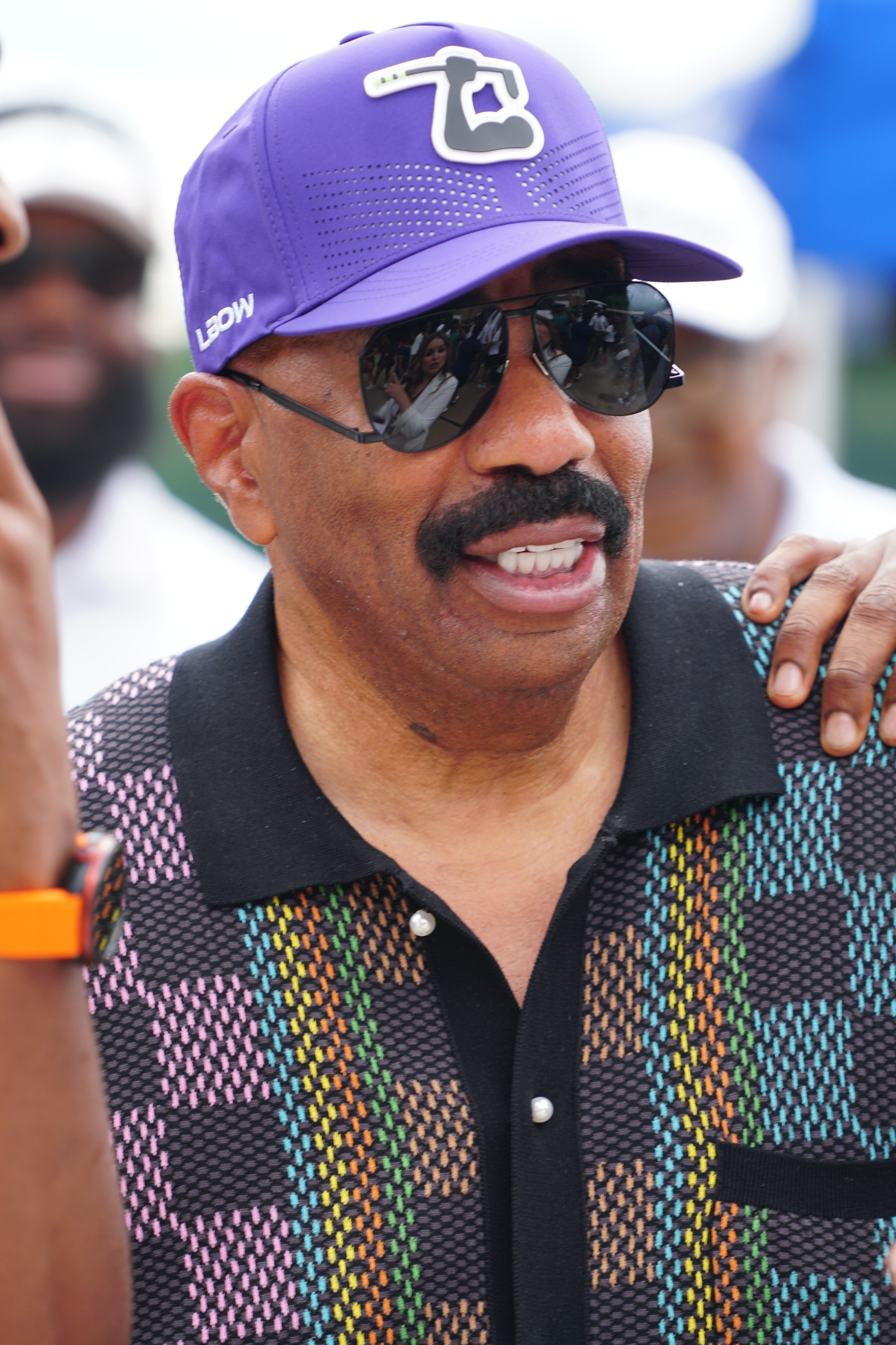 A smiling man wearing a purple baseball cap with a logo and sunglasses, standing in a crowd and wearing a colorful patterned shirt.