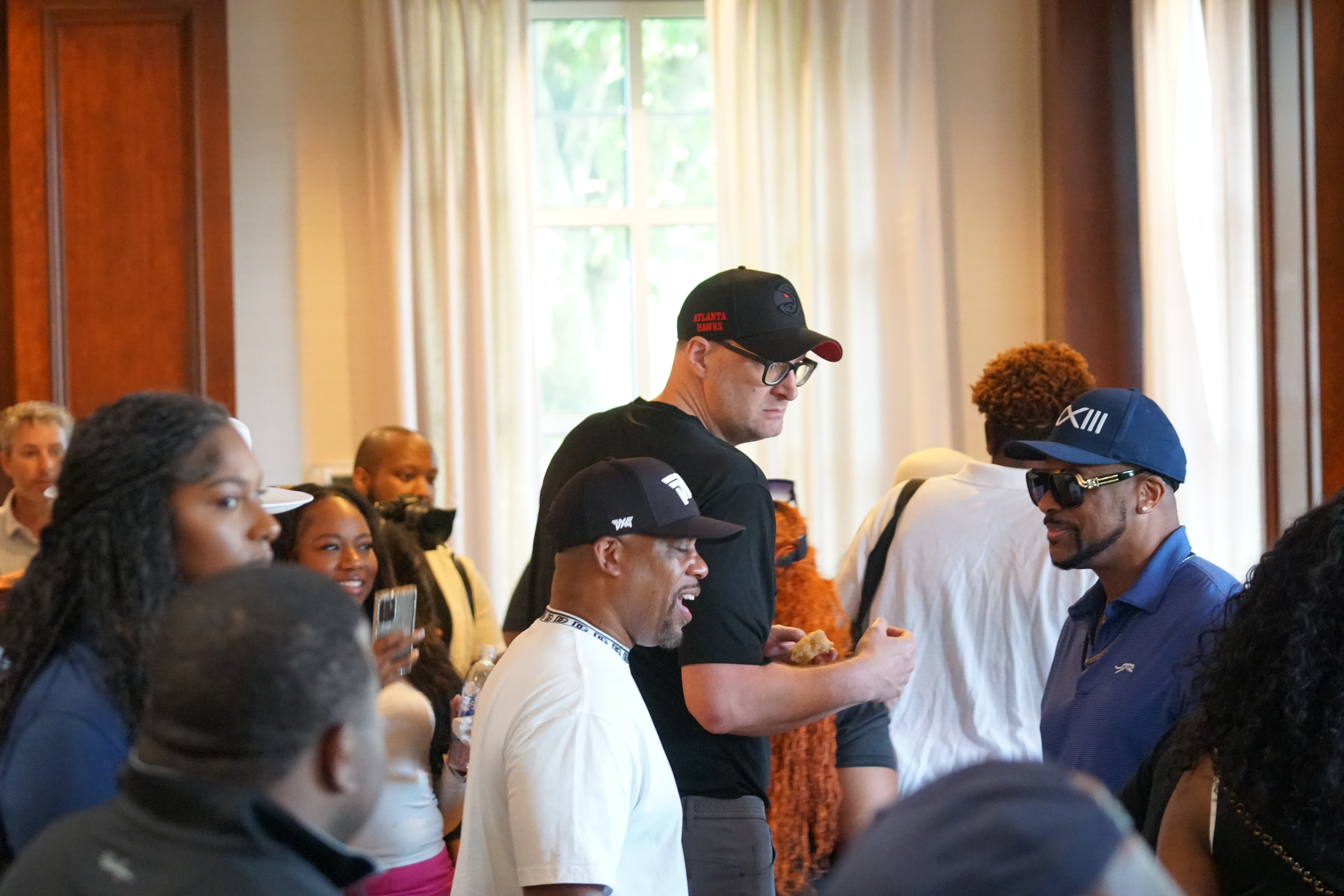 A group of people, some wearing hats and shirts with logos, gathered in an indoor setting with curtained windows.