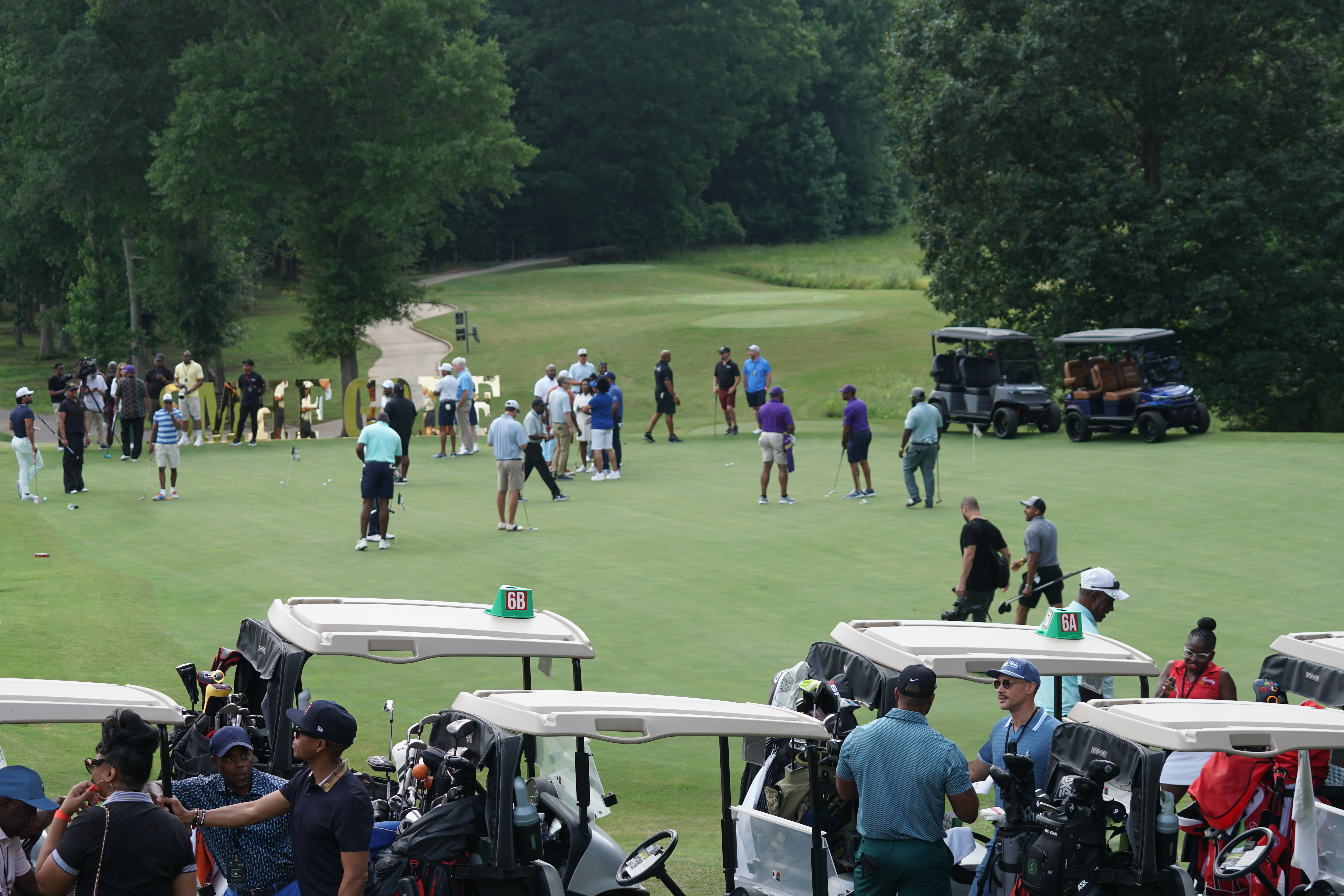 A golf course with a group of people playing on the green, surrounded by golf carts and spectators.