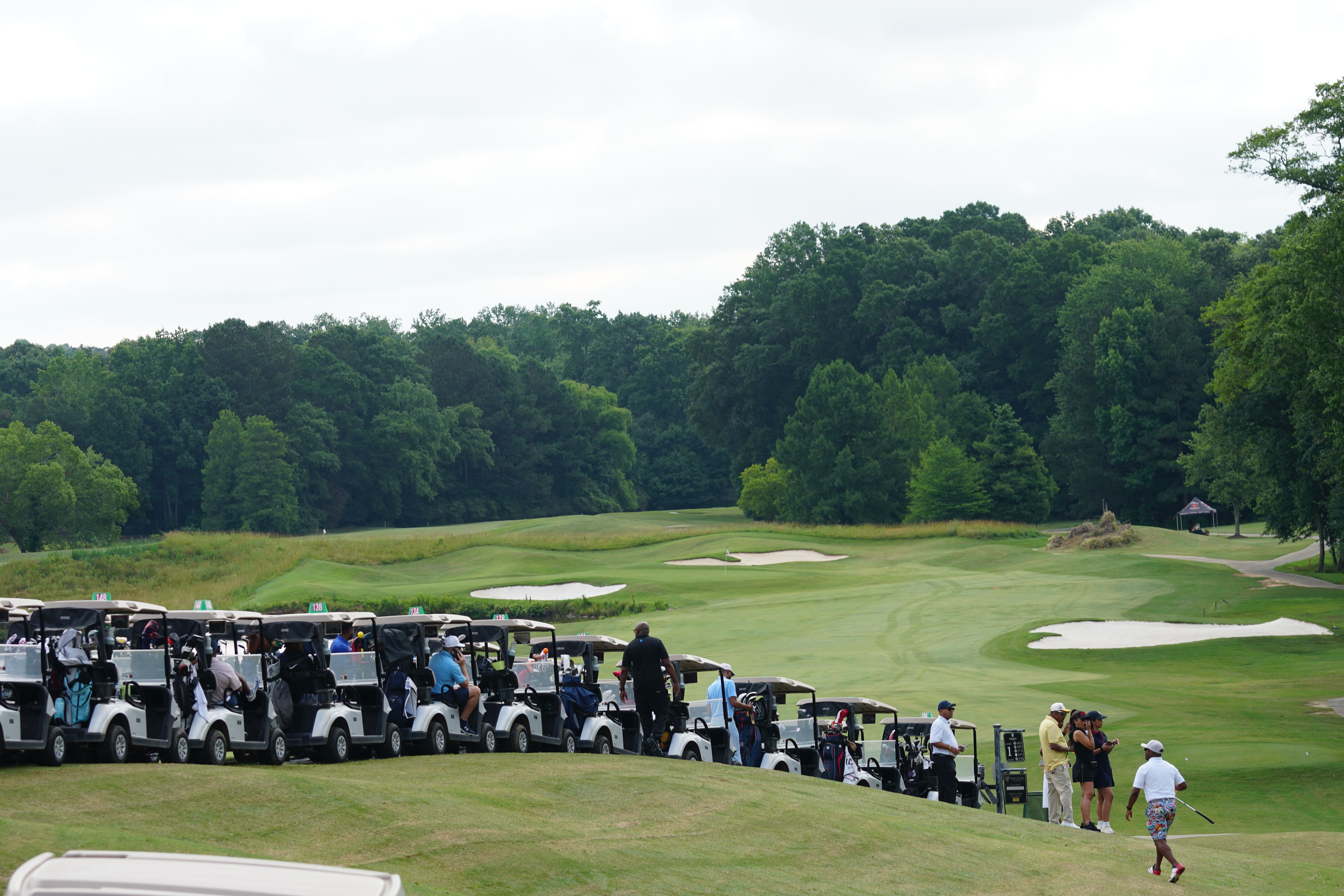 A golf course with a fairway surrounded by lush green trees. Several golf carts are parked along the edge of the fairway, and people can be seen walking on the course.
