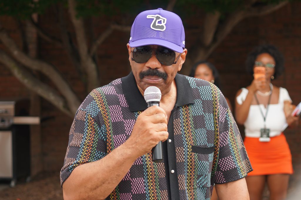 A man wearing a purple baseball cap with the number 73 and sunglasses, holding a microphone and speaking into it on a stage.
