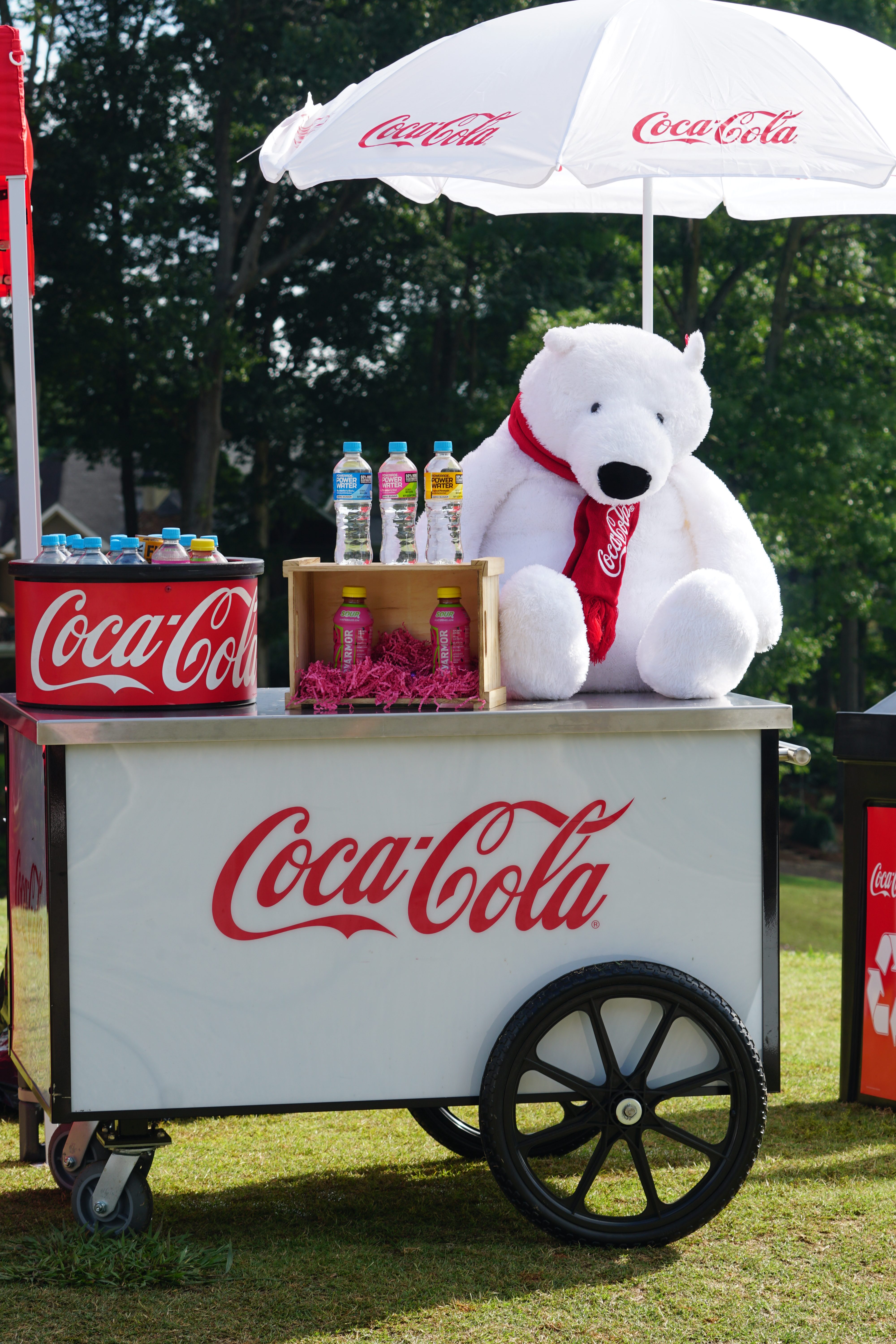 A Coca-Cola branded cart with a white plush bear, Coca-Cola branded umbrellas, and various Coca-Cola beverages on display in a grassy outdoor setting.