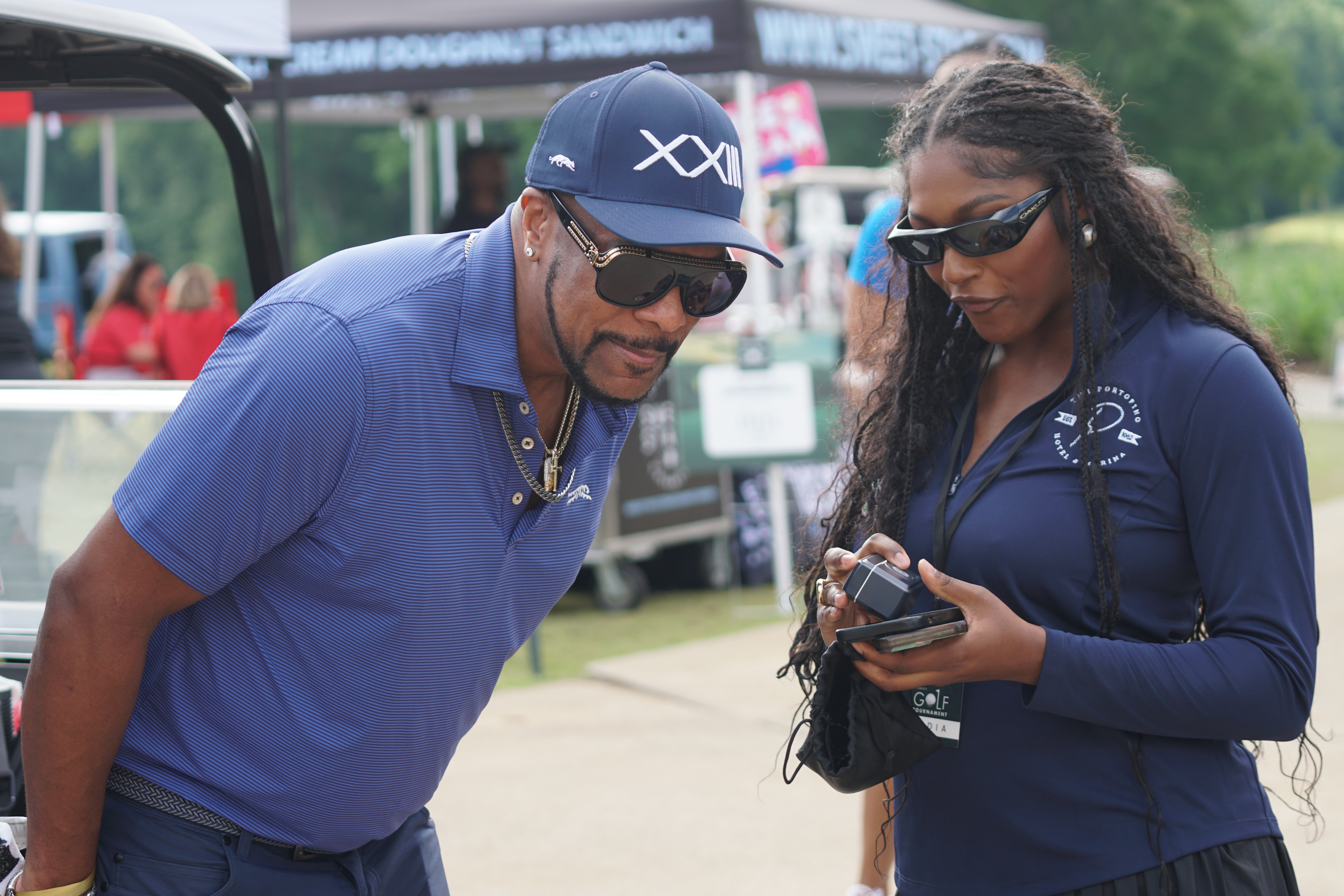 Two people wearing sunglasses and branded clothing standing near a golf cart and event tents.