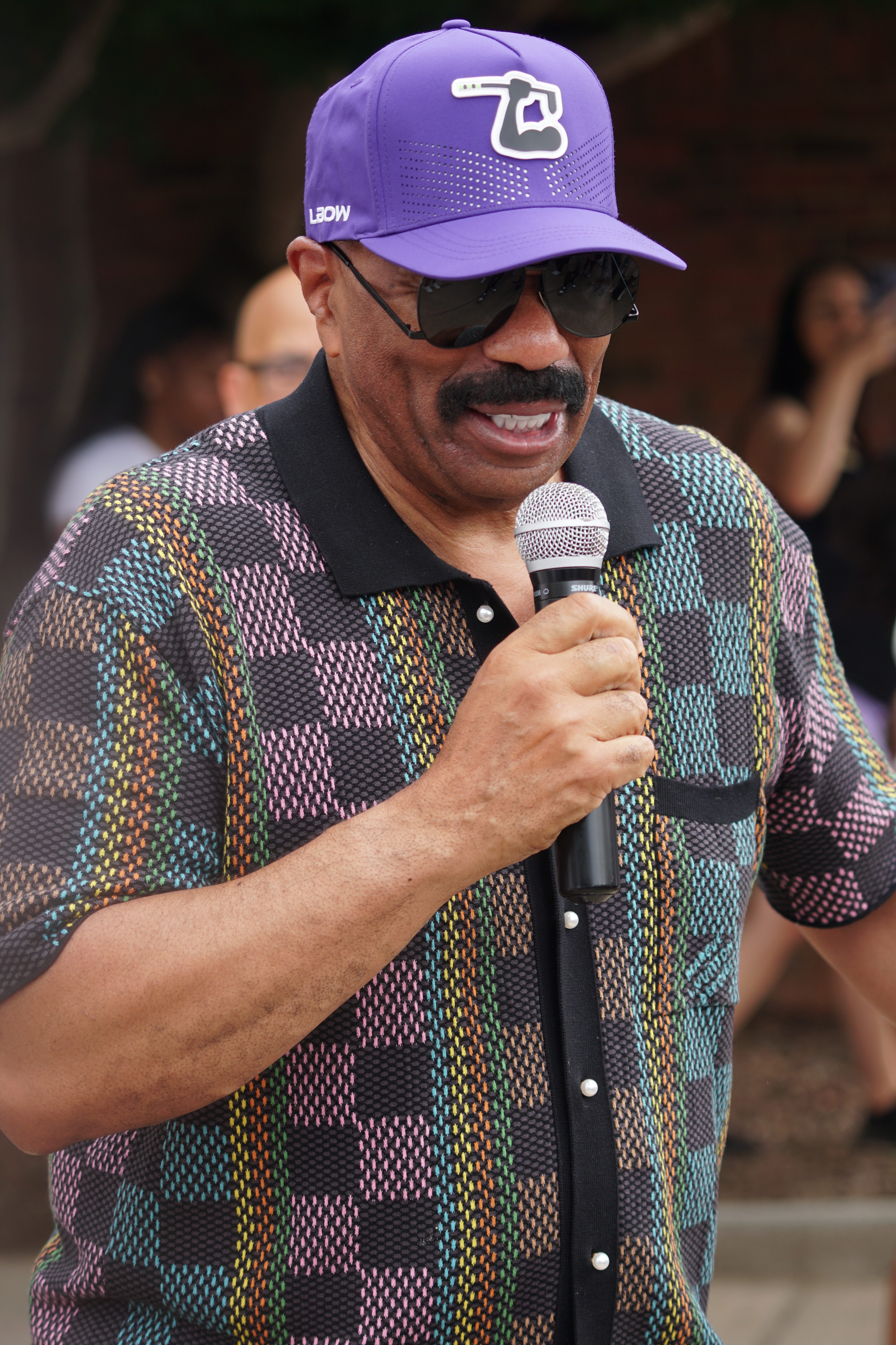 A man wearing a purple baseball cap with the logo "76" and a colorful patterned shirt holding a microphone and smiling.