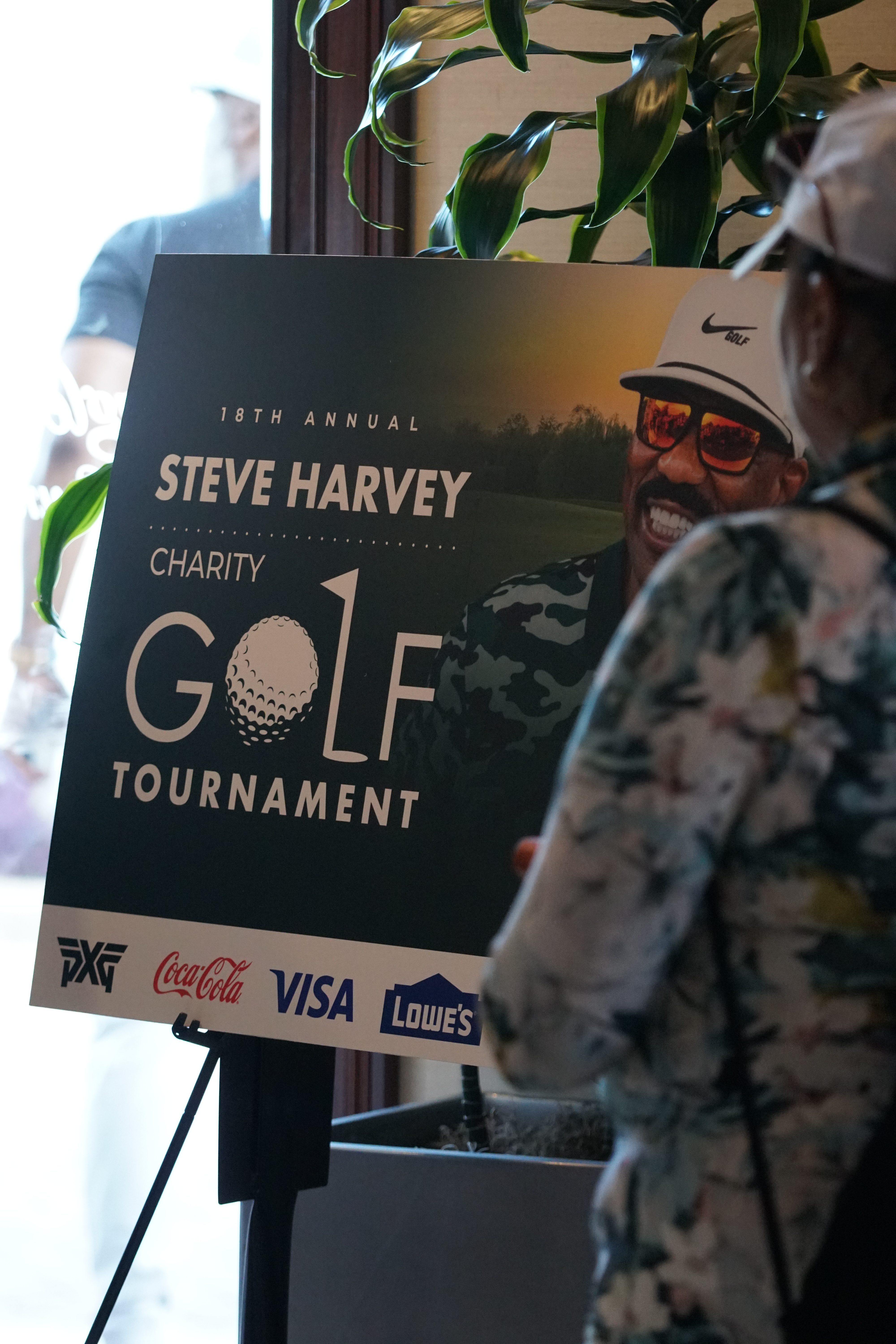 18th Annual Steve Harvey Charity Golf Tournament banner featuring a smiling man in sunglasses, with logos for PXG, Coca-Cola, Visa, and Lowe's.