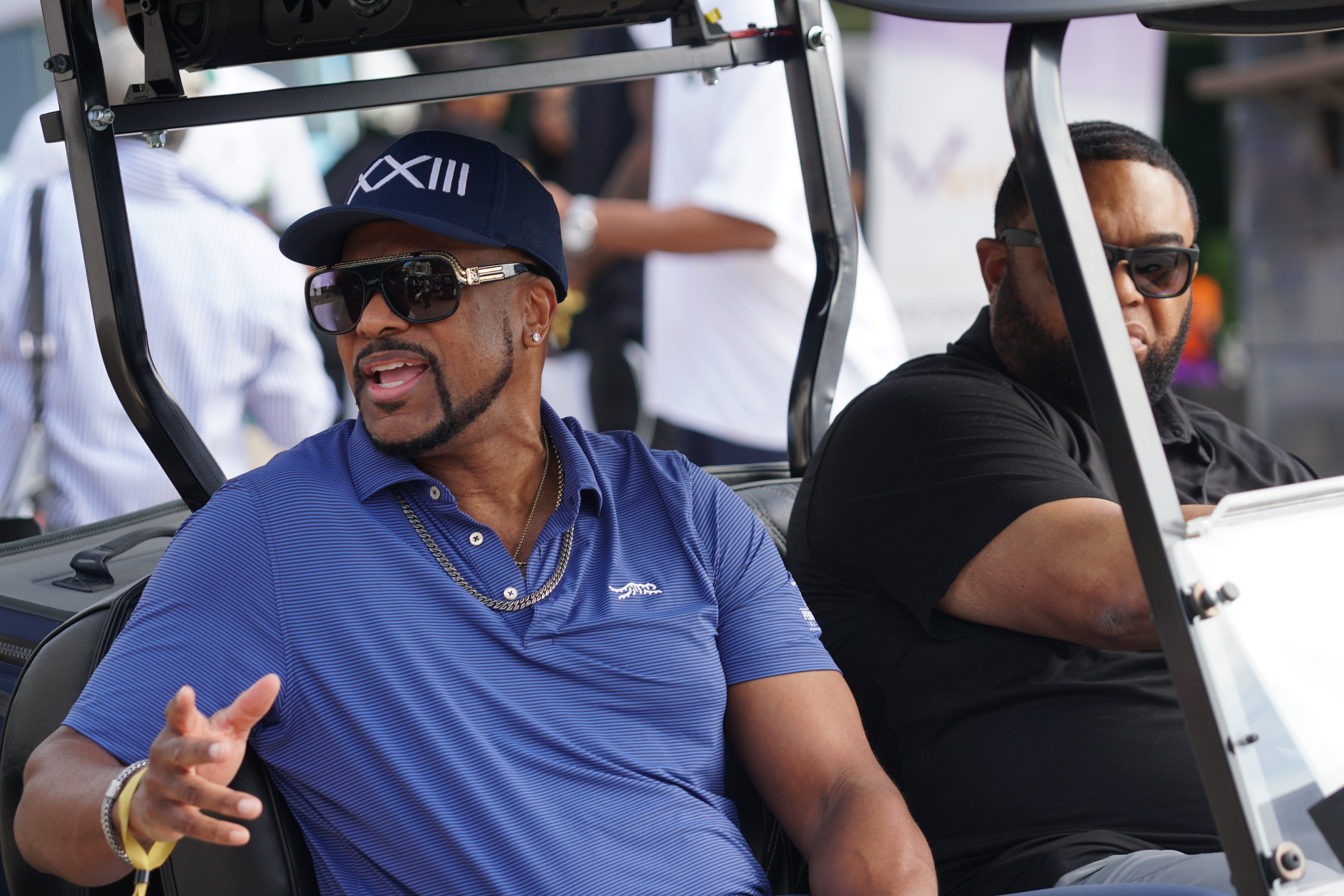 Two men, one wearing a blue polo shirt and baseball cap with the number 13, and the other wearing a black shirt, sitting in a golf cart.