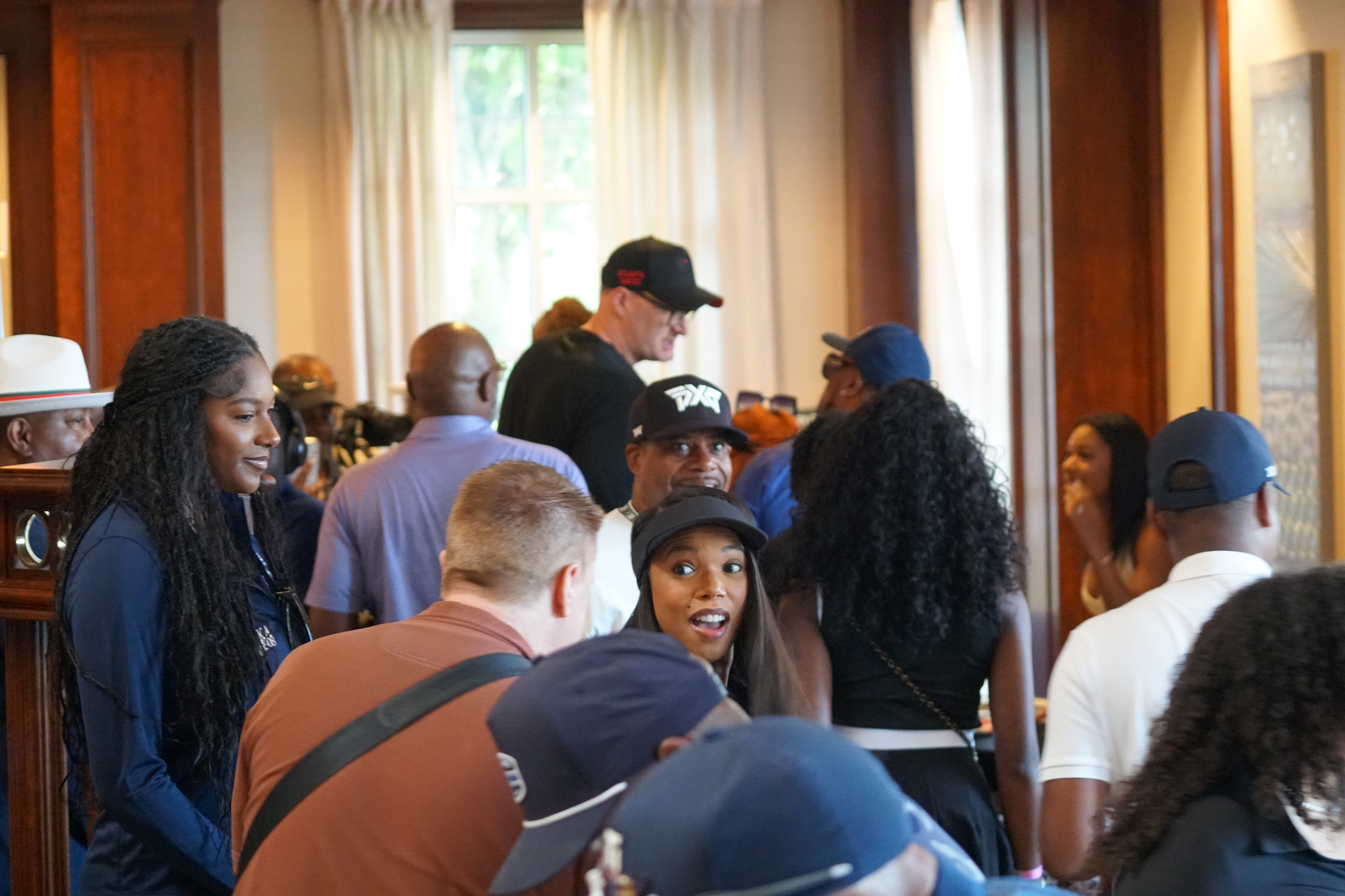 A group of people, mostly young adults, gathered in what appears to be a lobby or common area. Several are wearing hats and casual clothing, and some are smiling or engaged in conversation.
