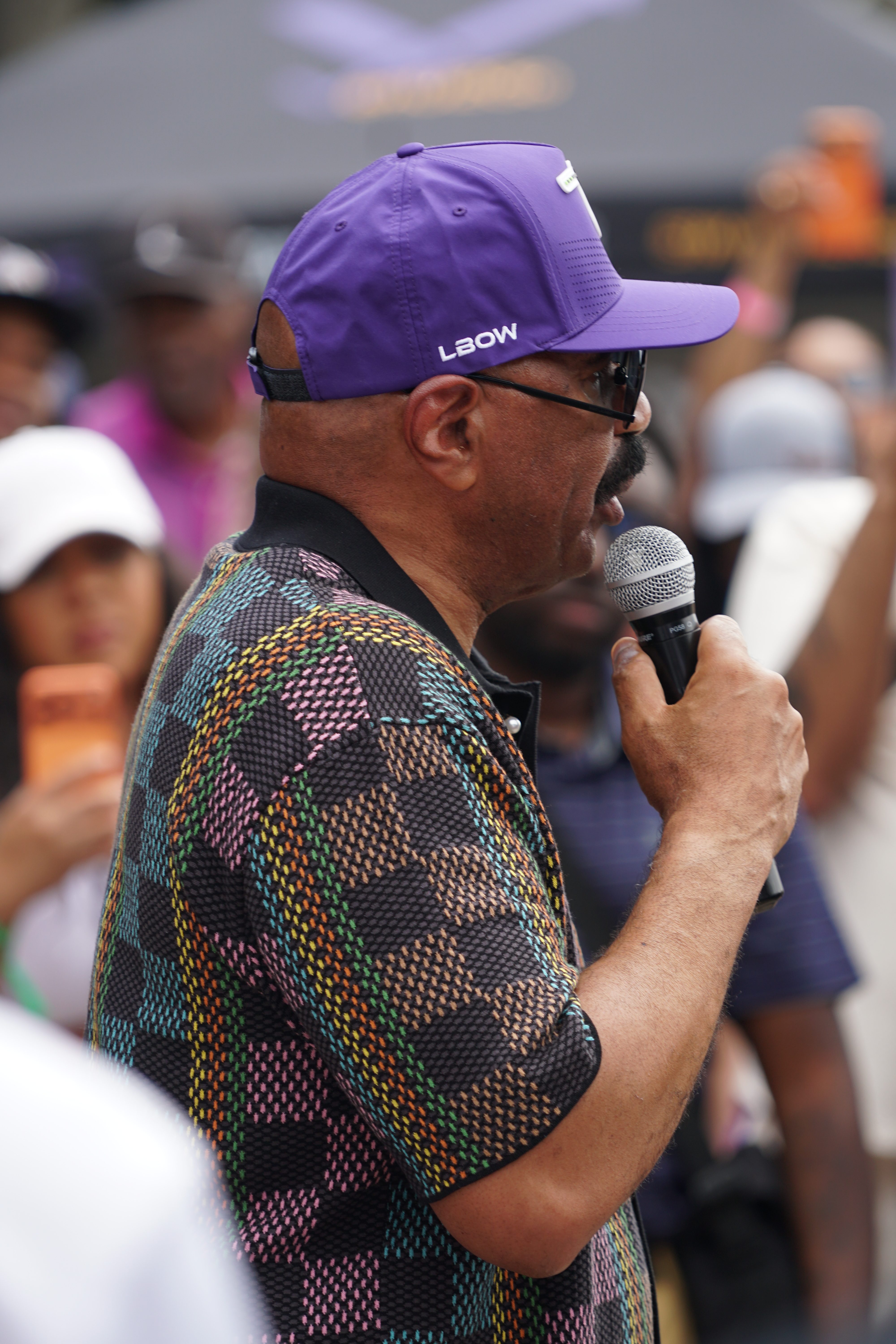 A person wearing a purple LBOW cap and a colorful patterned shirt speaking into a microphone at an event.