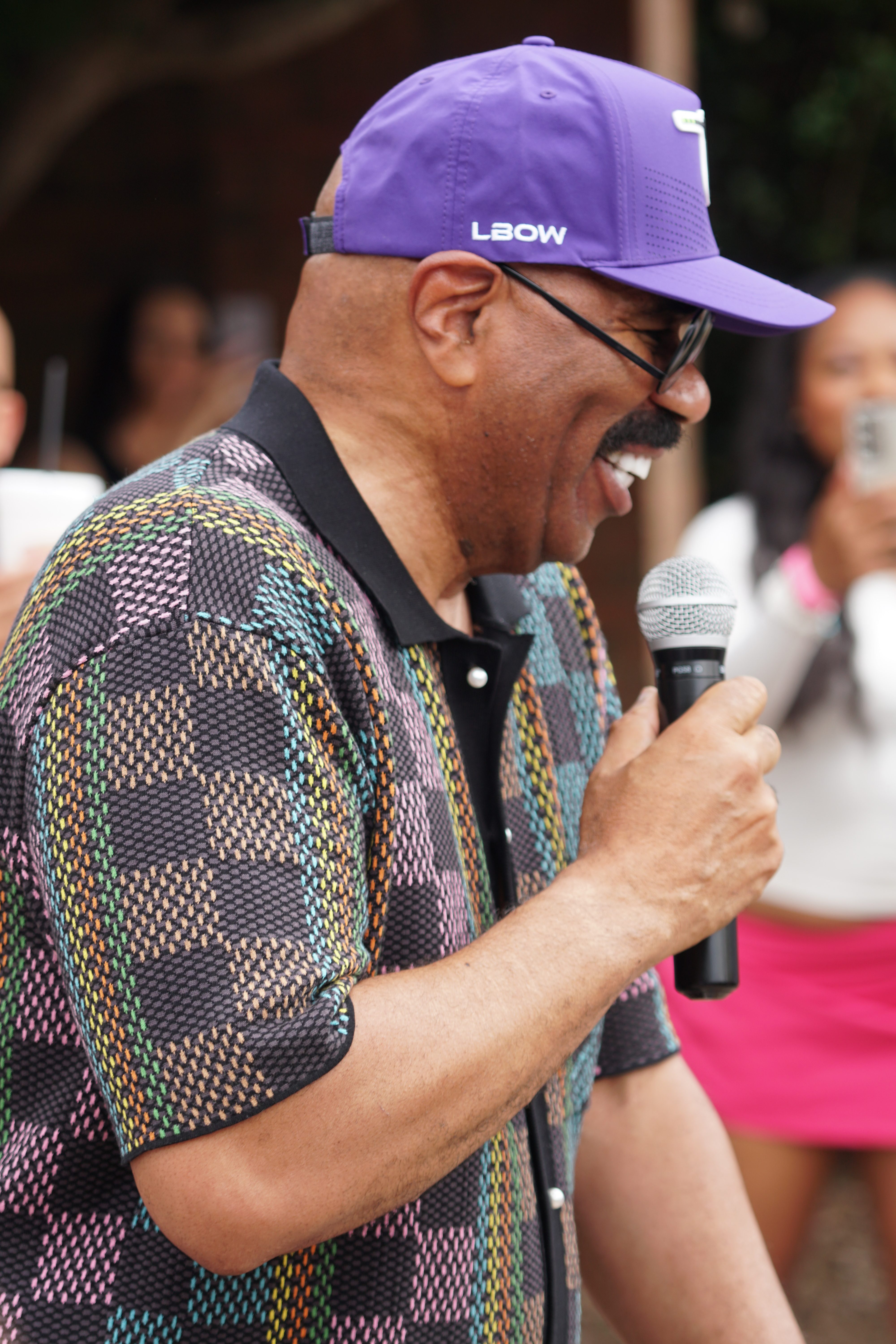 A man wearing a purple cap with "LBOW" text and a colorful patterned shirt speaking into a microphone.