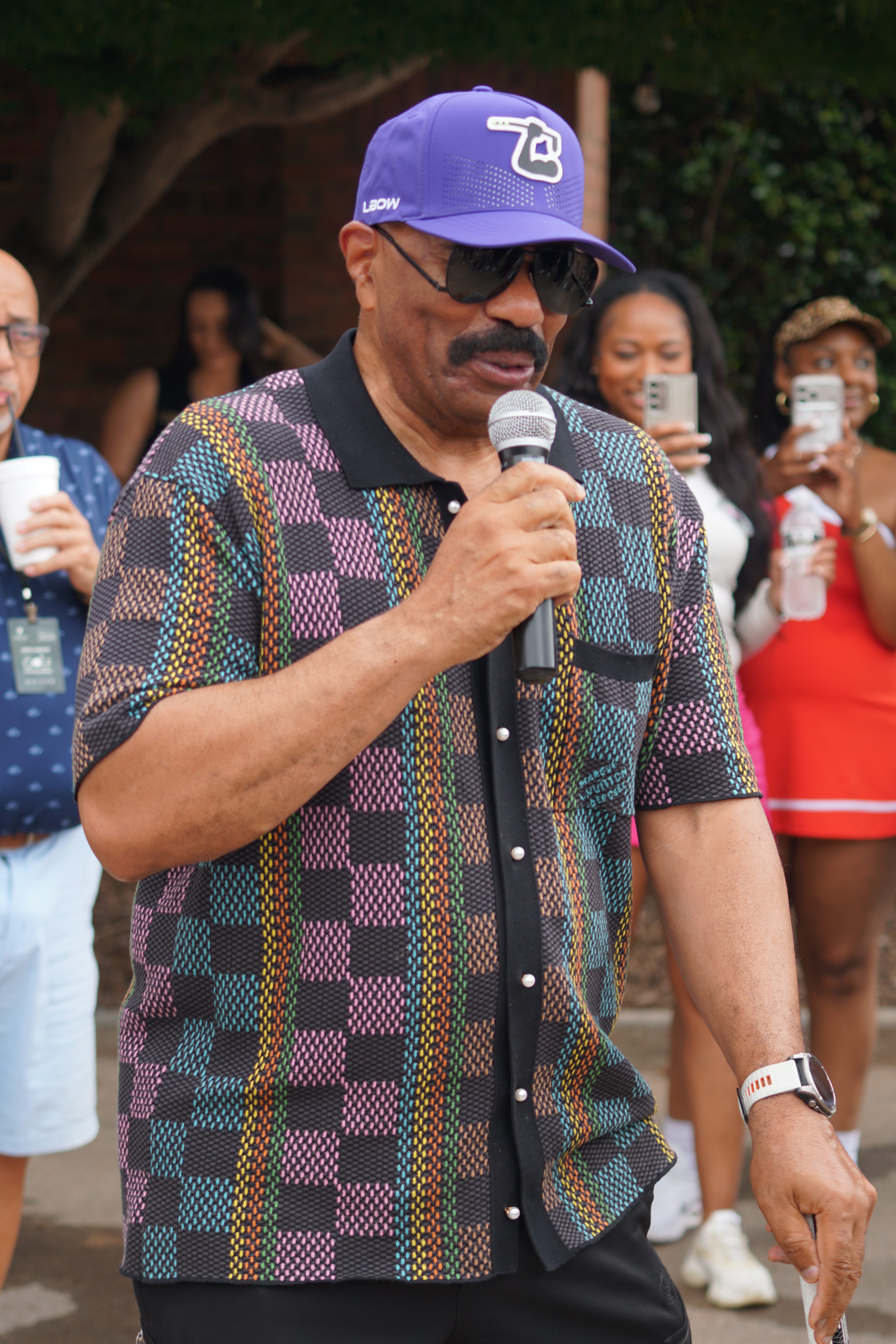 A man wearing a colorful patterned shirt and a purple baseball cap with a logo, holding a microphone and speaking at an event.