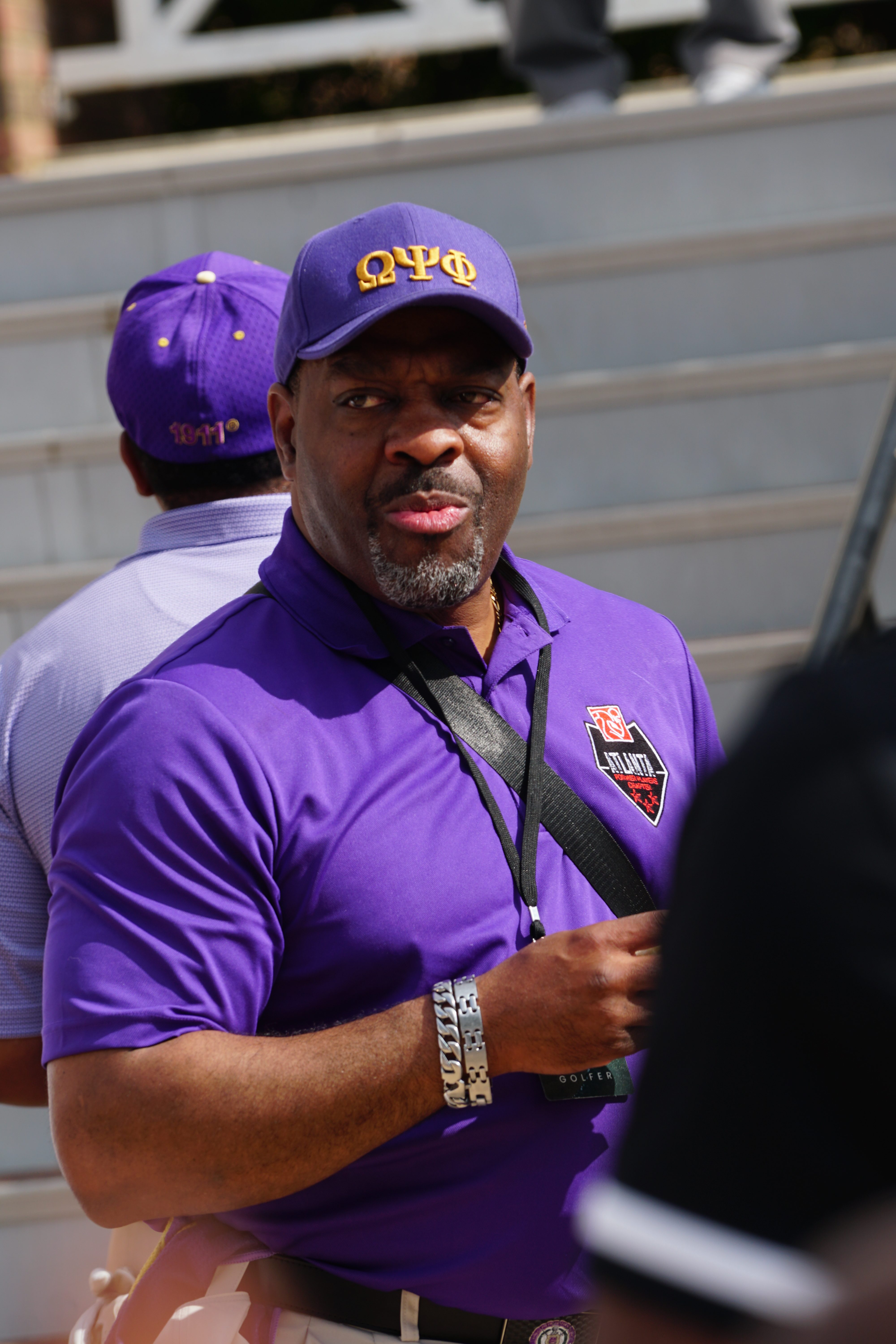 A middle-aged Black man wearing a purple shirt and cap with the Greek letters Omega Psi Phi, standing outdoors.