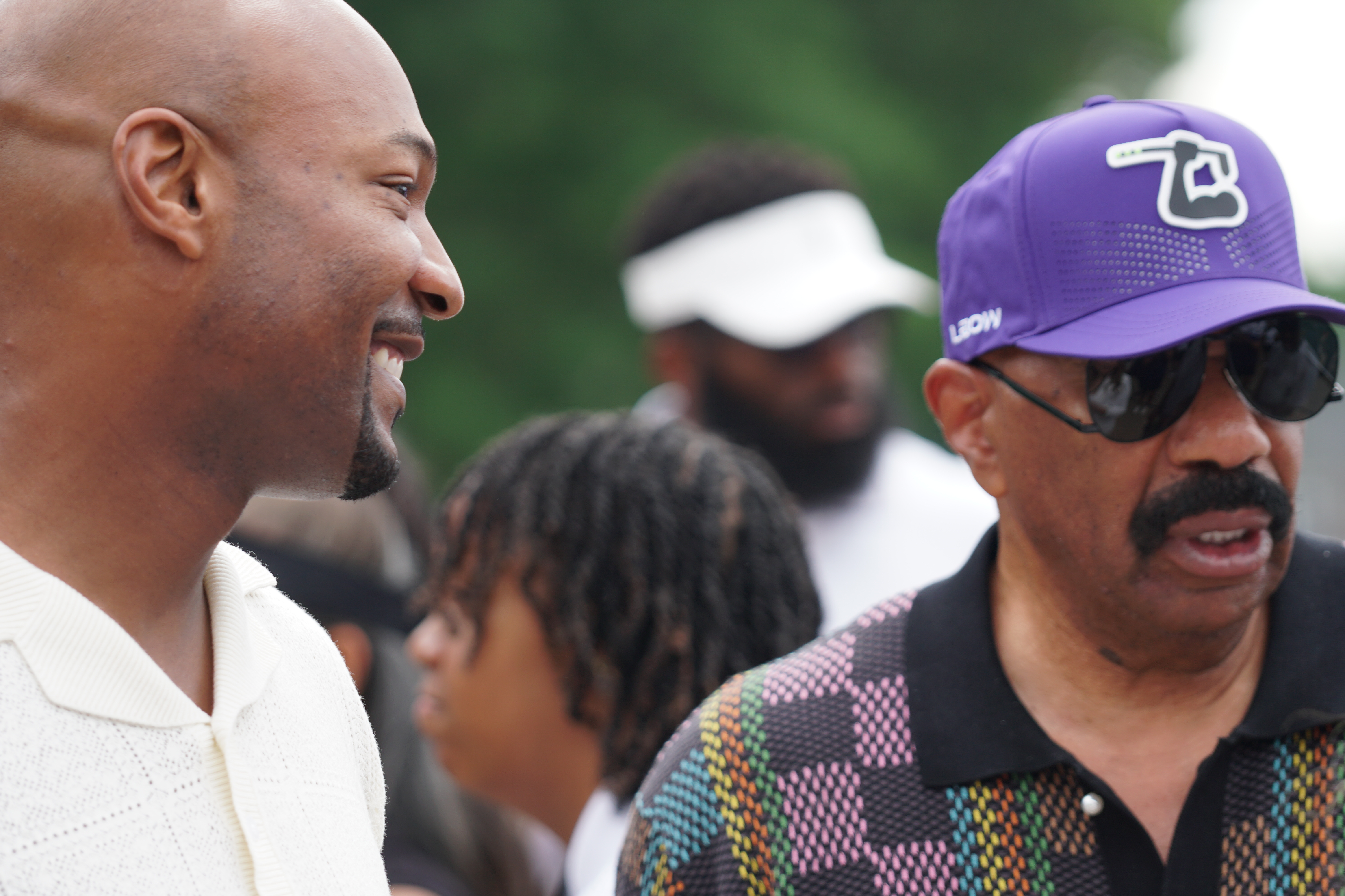 Two men in casual clothing, one wearing a purple baseball cap, engaged in conversation outdoors.