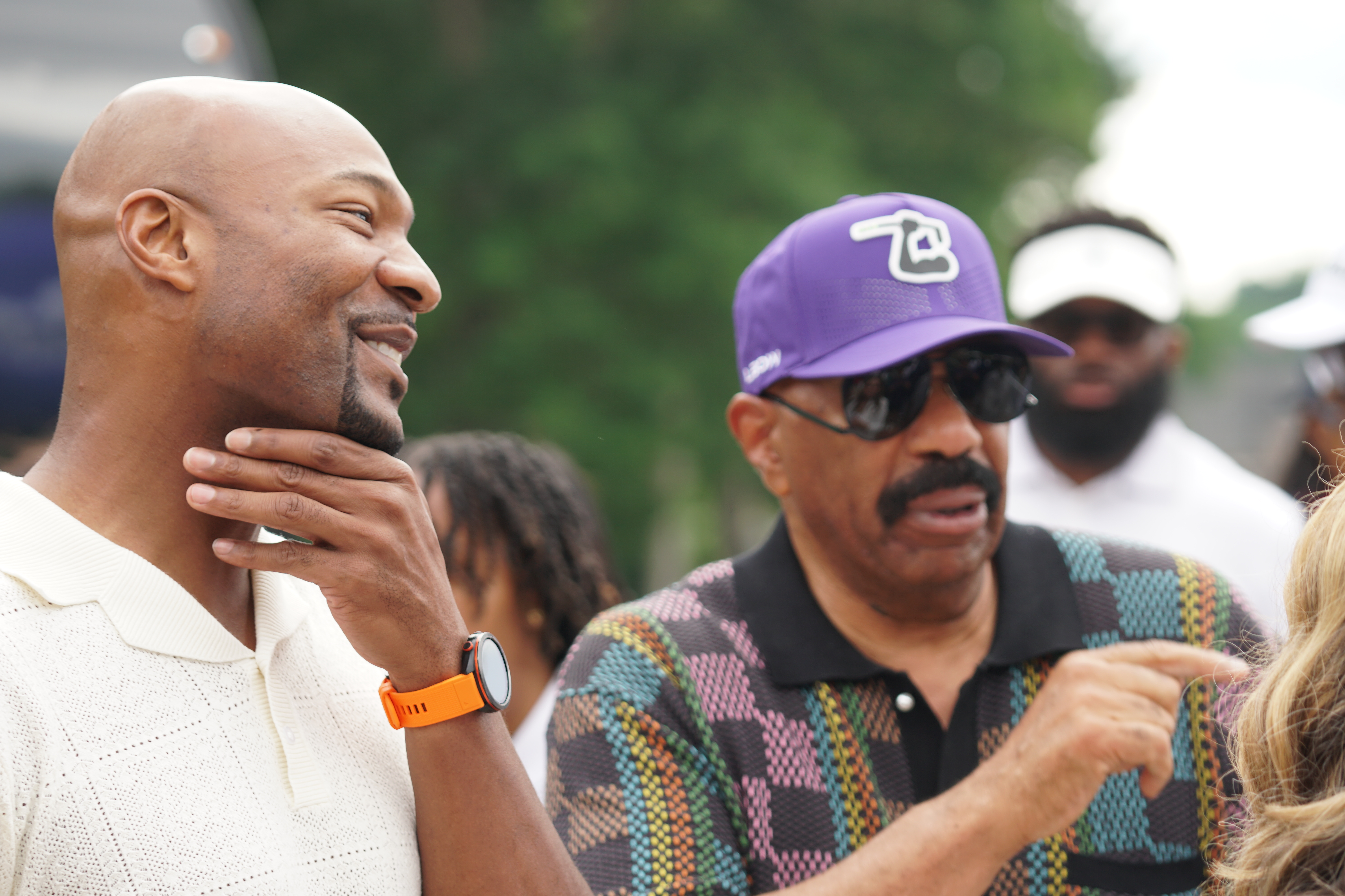 Two men in casual clothing engaged in conversation, one wearing a purple baseball cap with a logo.