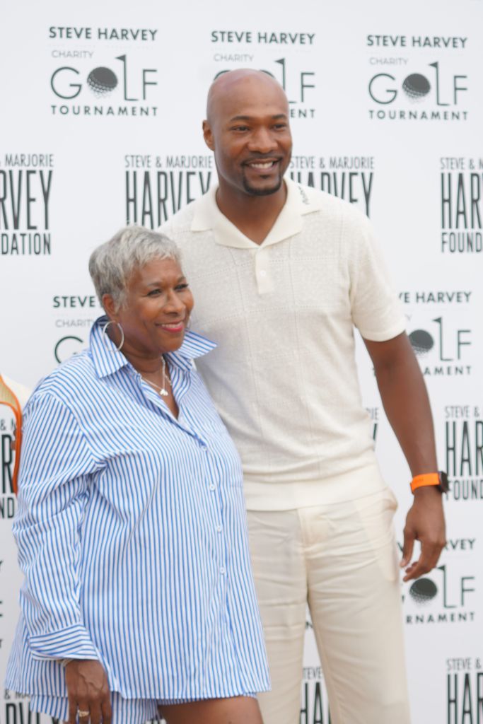 Two people, a smiling older woman and a taller man, standing in front of a backdrop with "Steve Harvey Charity Golf Tournament" text.