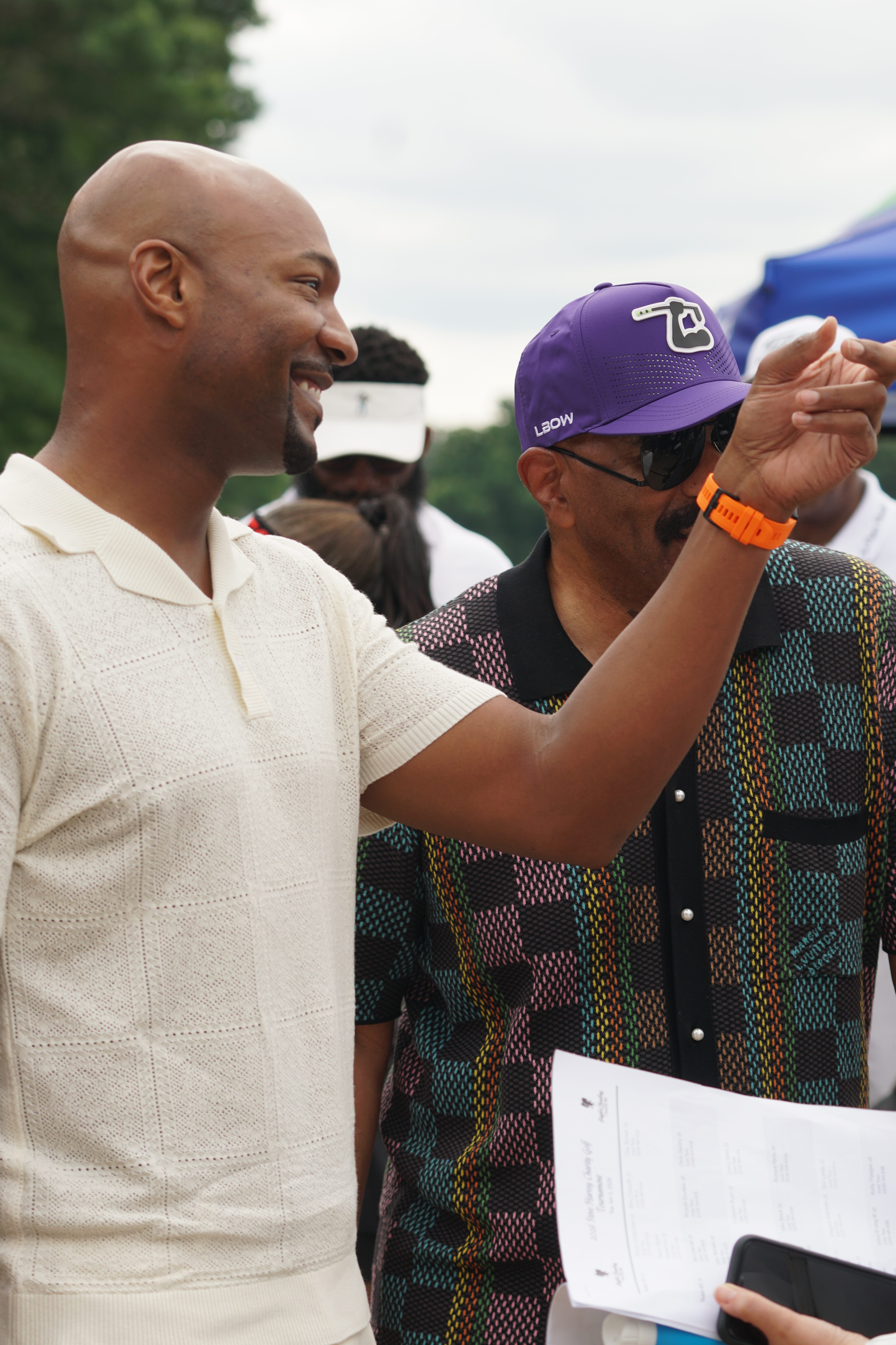Two men, one wearing a purple hat with "LBD" logo, interacting and smiling in an outdoor setting.