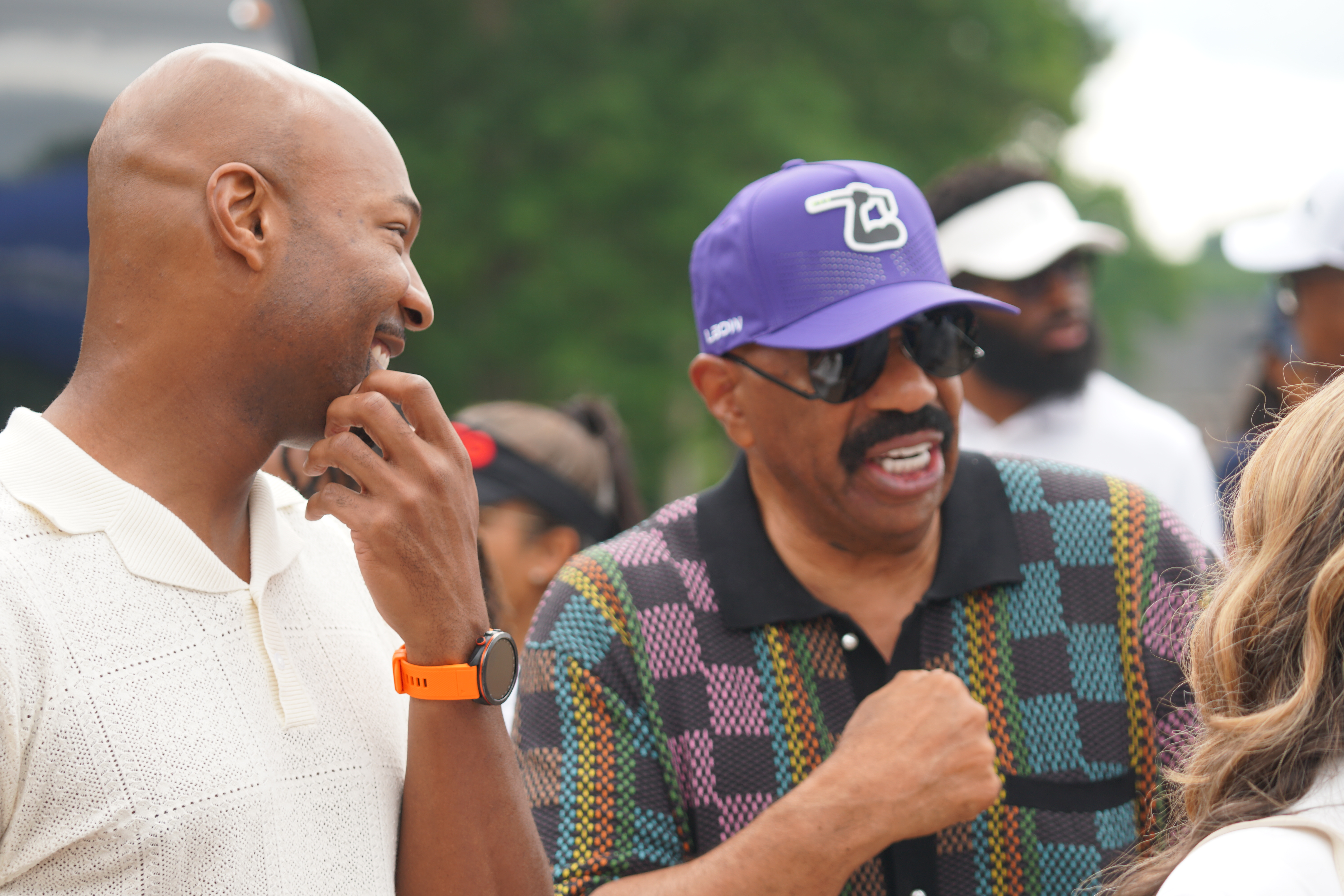 Two men in casual clothing engaged in conversation, one wearing a purple baseball cap with a logo.