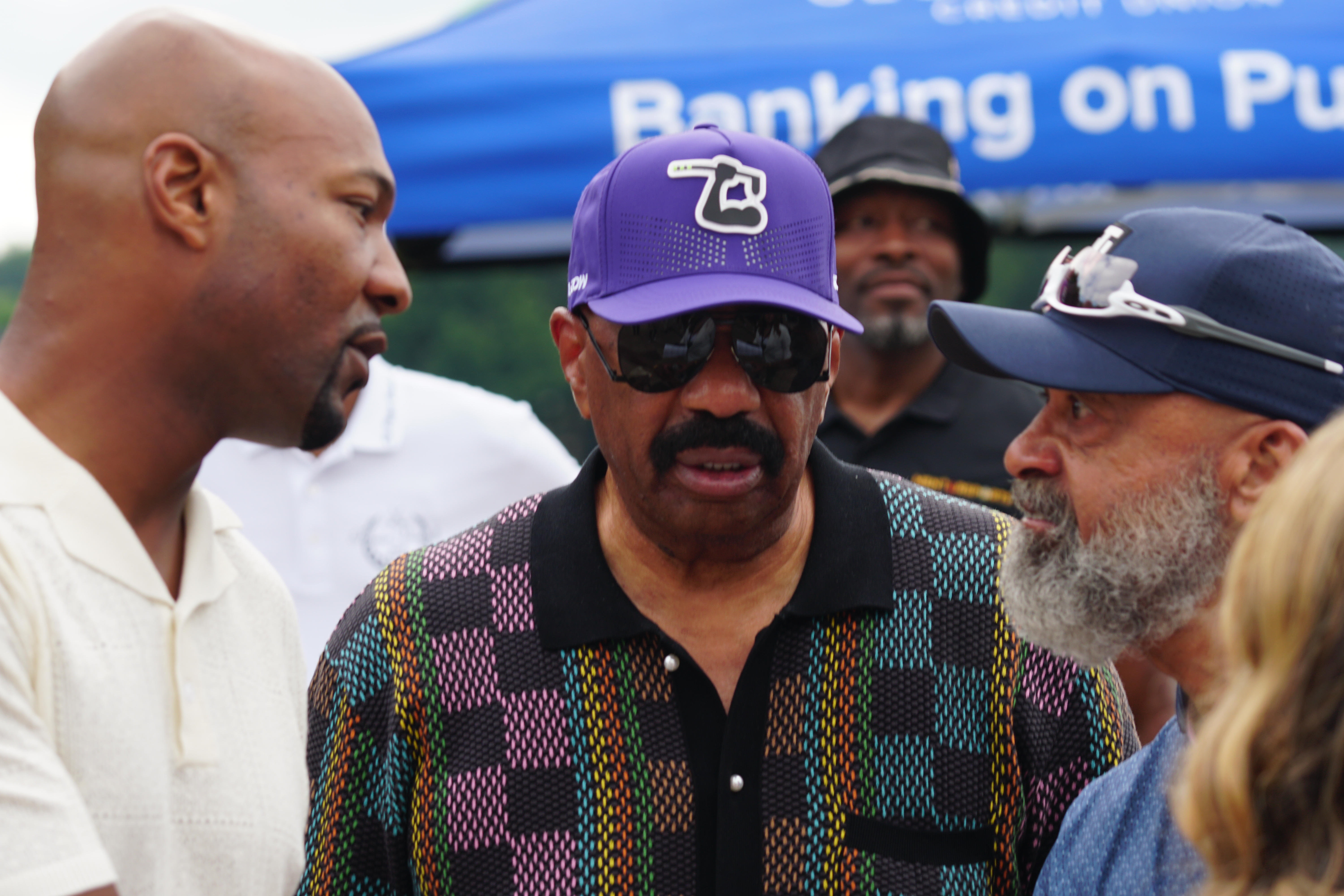 Three men, one wearing a purple hat with a logo, standing in front of a blue tent with the text "Banking on Pu".