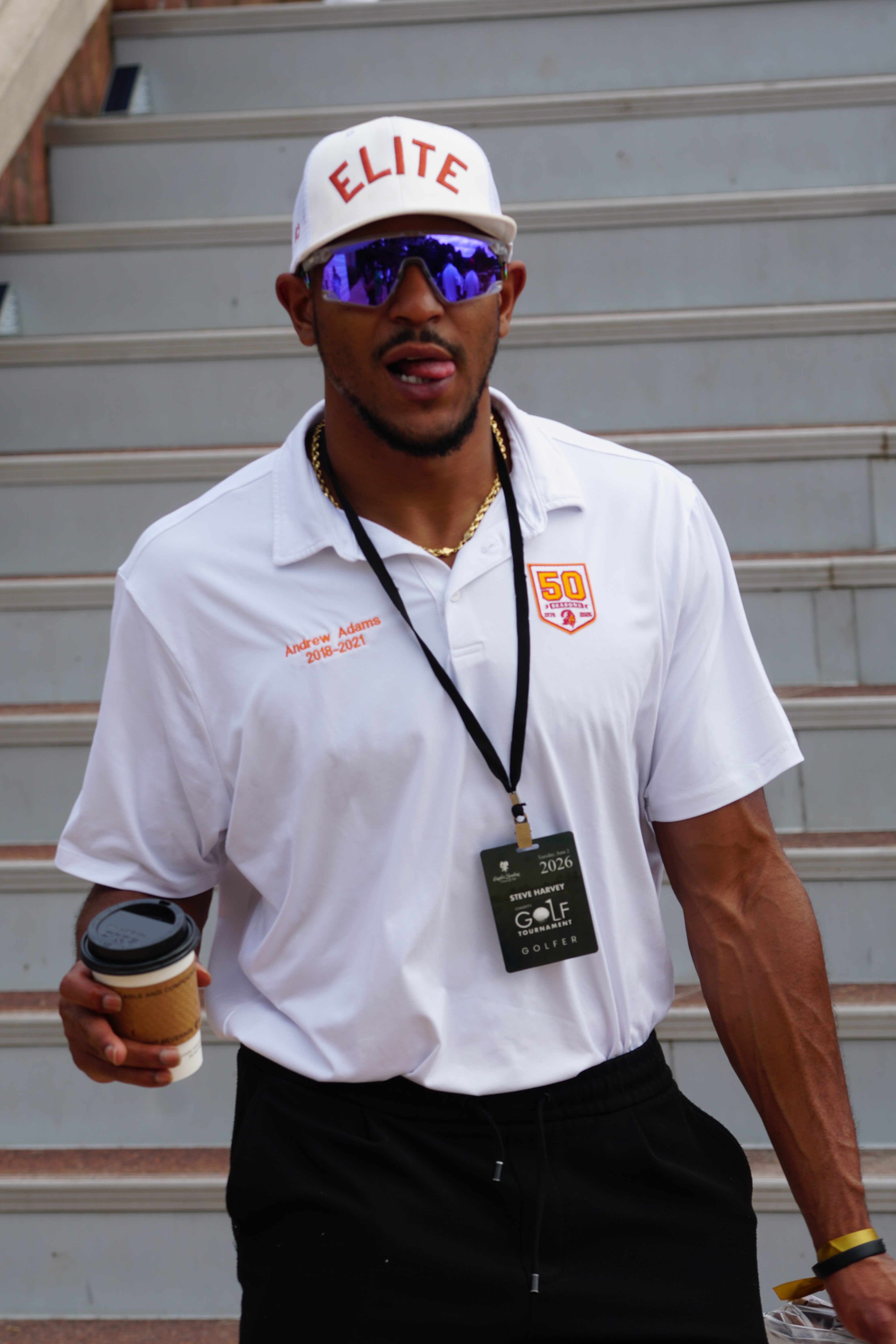 A man wearing a white "Elite" hat and sunglasses, holding a coffee cup, with a "2026 GOLF" credential around his neck.