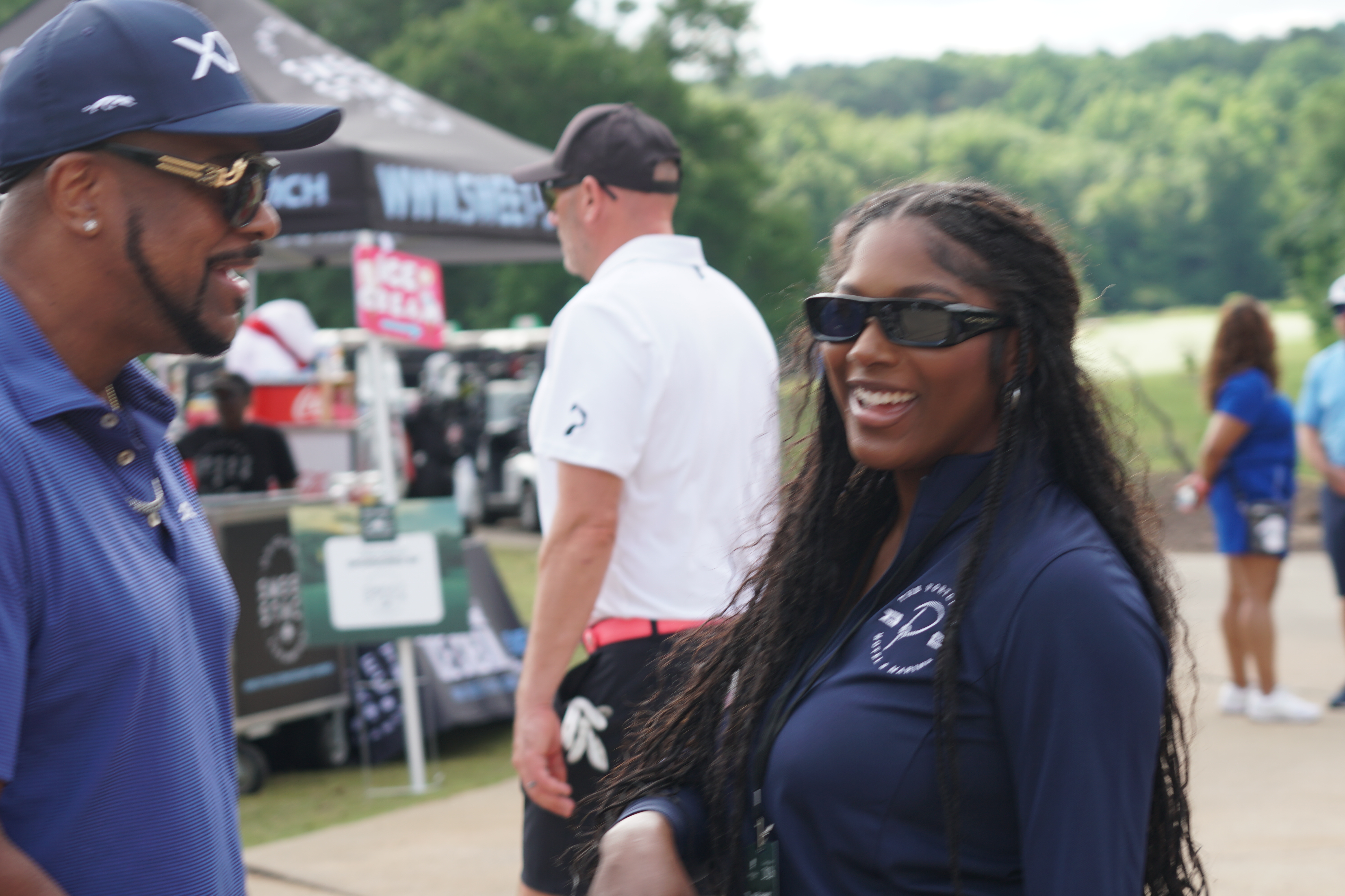 Two people, a man and a woman, smiling and talking at an outdoor event with tents and booths in the background.