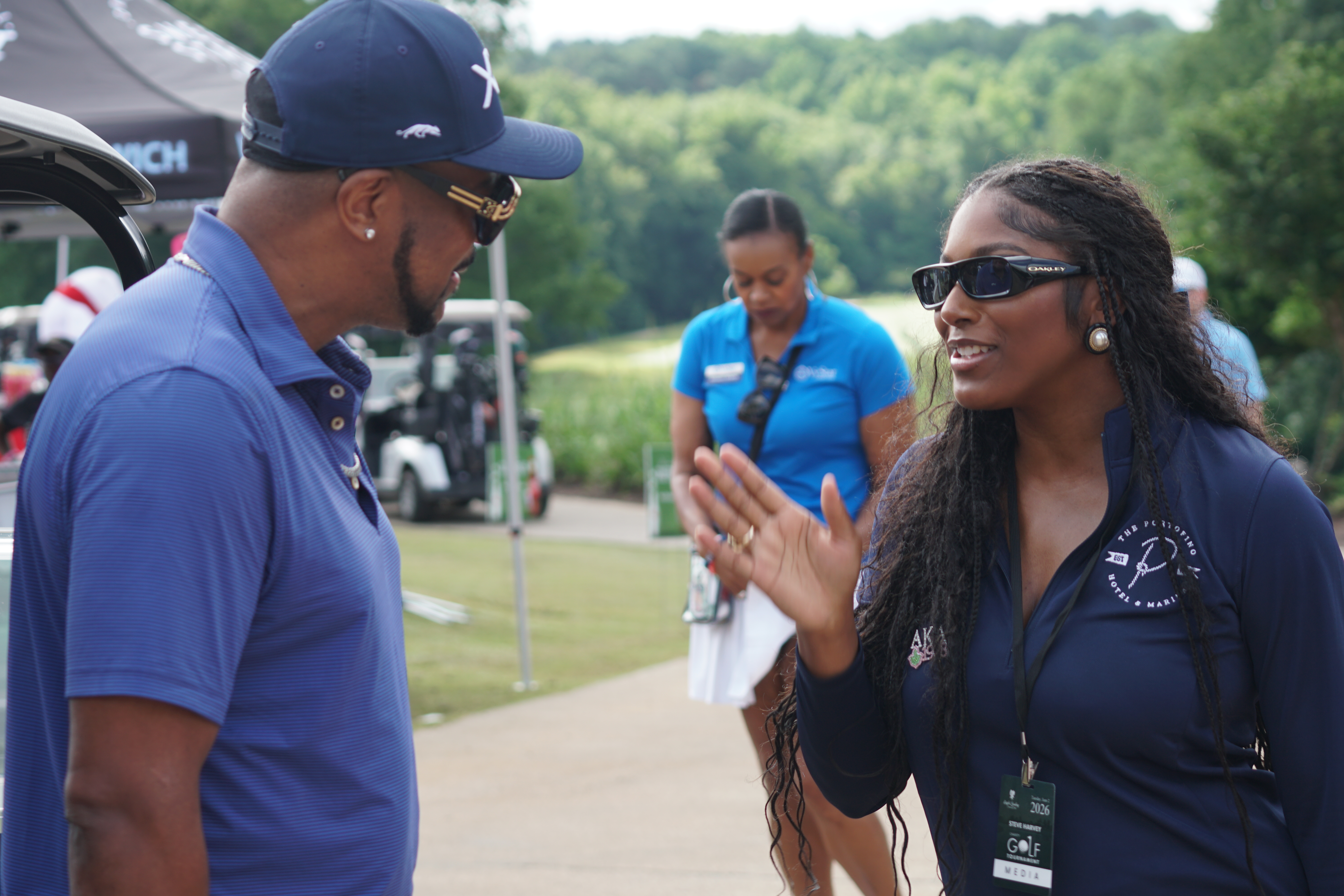 Two people in sports uniforms conversing on a golf course with trees in the background.